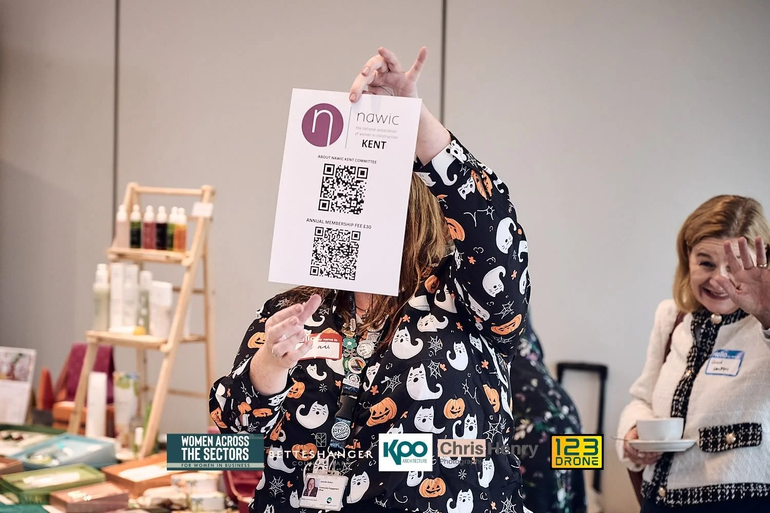 Woman in Halloween-themed sweater holding a paper with QR codes in front of her face, women smiling and talking at an indoor event with display shelves and branded signage.