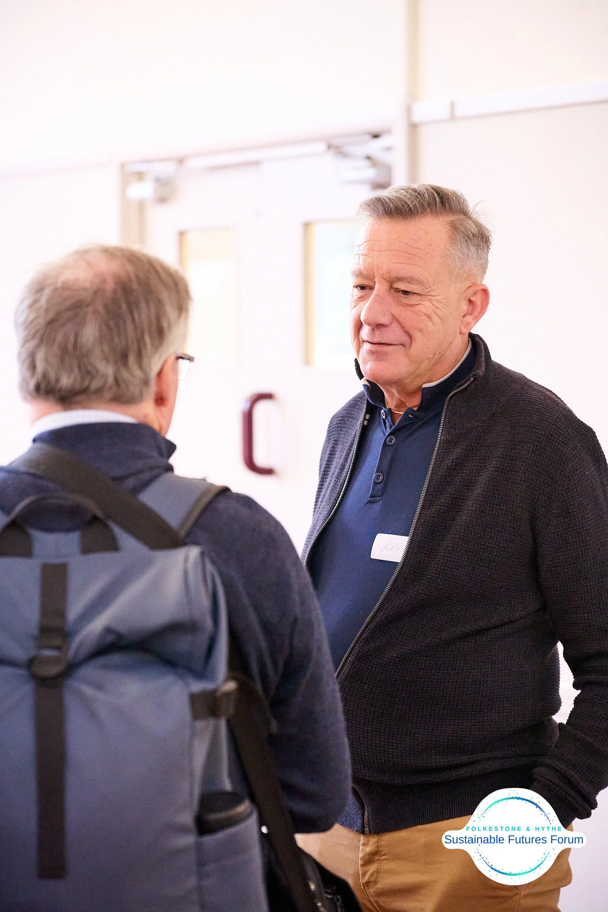 Two men engaged in conversation at the Folkestone & Hythe Sustainable Futures Forum, one with a backpack and the other wearing a dark jacket and name tag.