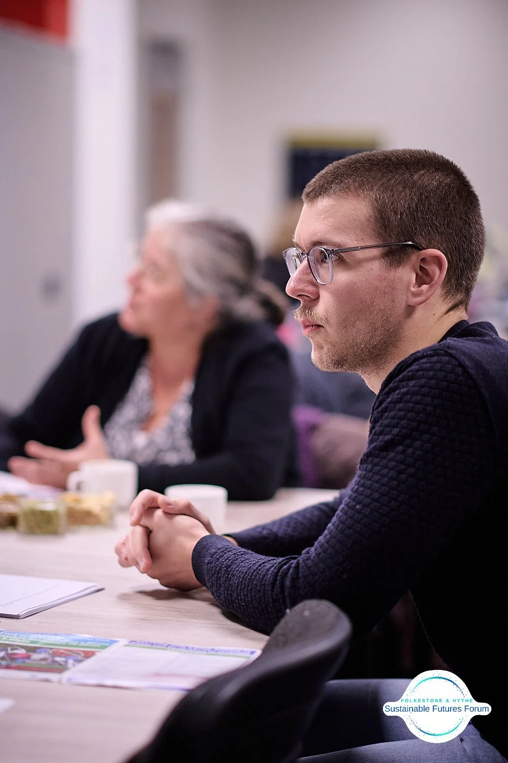 A young man with glasses sits at a table during a meeting, with an older woman with gray hair next to him. Both are focused and engaged, with papers and cups on the table, at a Sustainable Futures Forum event.
