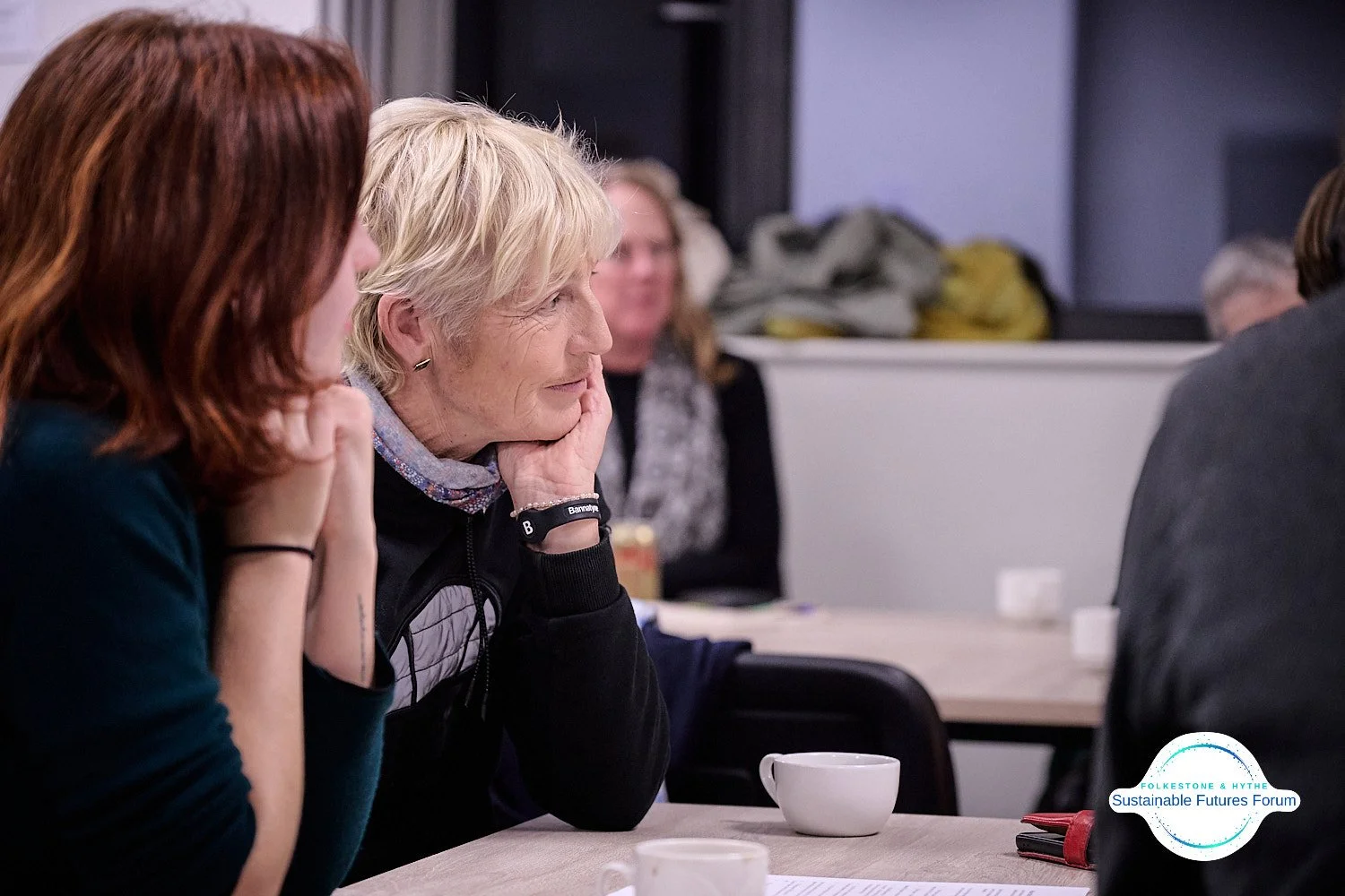 A group of people sitting at a table during a meeting or discussion, with a woman with short blonde hair resting her chin on her hand, listening attentively.