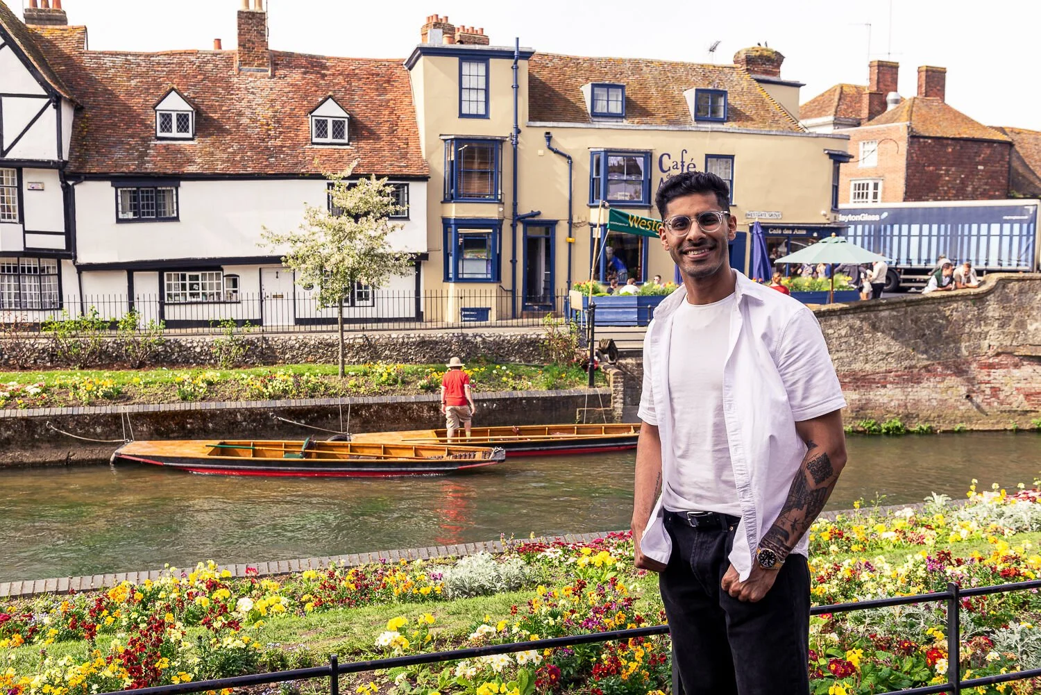 A smiling man with tattoos, glasses, and a white shirt over a t-shirt standing near a garden and canal, with a boat and historic buildings in the background.