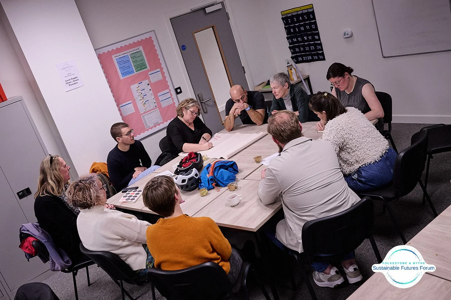 A group of twelve people sitting around a rectangular table in a meeting room, engaged in a discussion. Items on the table include a bicycle helmet, backpacks, notebooks, and small bowls. The room has a whiteboard, a clothing calendar, and pink bulle