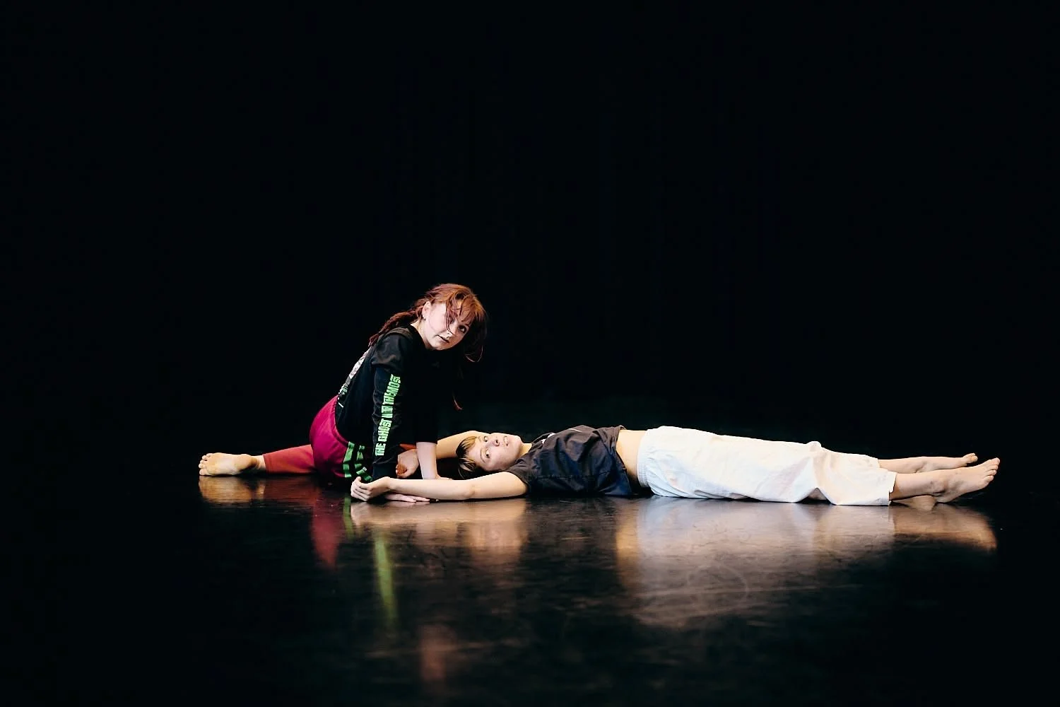 Two young women practicing dance on a black stage, one lying down and the other kneeling, both looking at the camera.