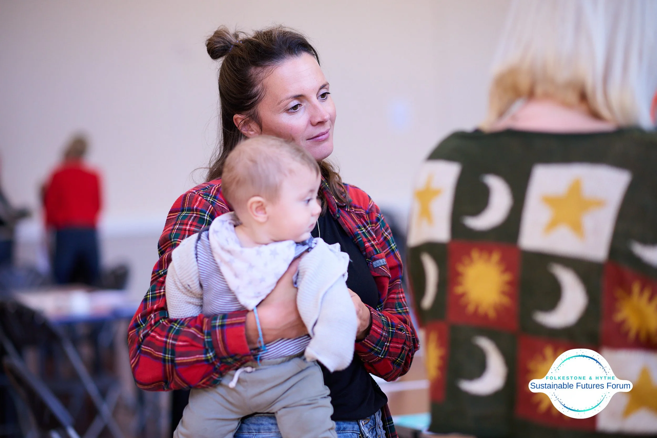 A woman holding a young child, observing a colorful display with a design featuring stars, moons, and suns, at the Folkestone & Hythe Sustainable Futures Forum.