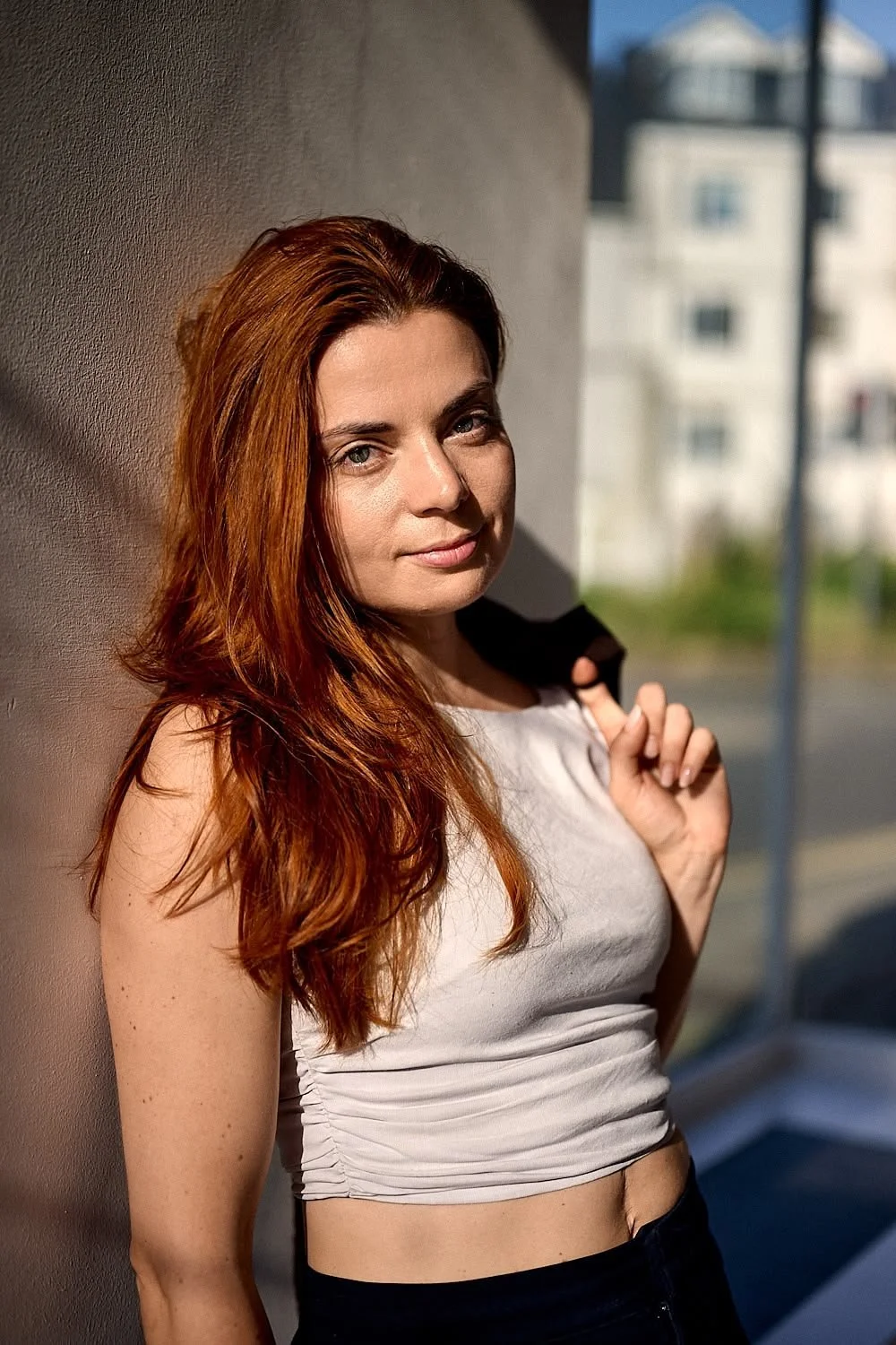 A young woman with long red hair and light skin standing indoors near a window, with sunlight illuminating her face. She wears a sleeveless white top and holds a black jacket over her shoulder, with a cityscape visible outside the window.