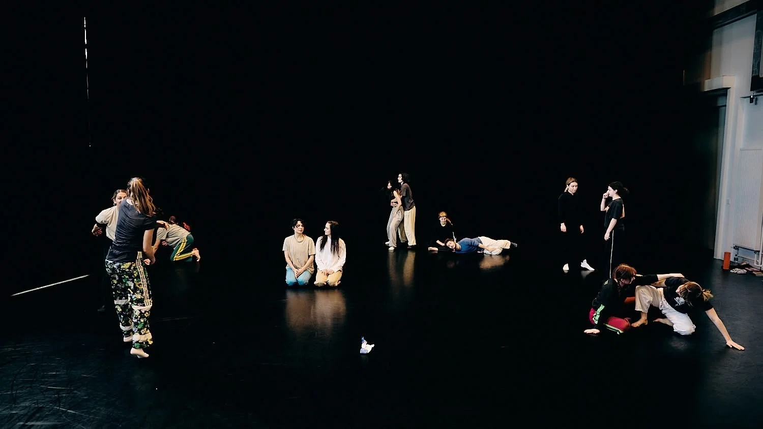 Group of people rehearsing or performing on a black stage with a plain black background, in various positions and actions.