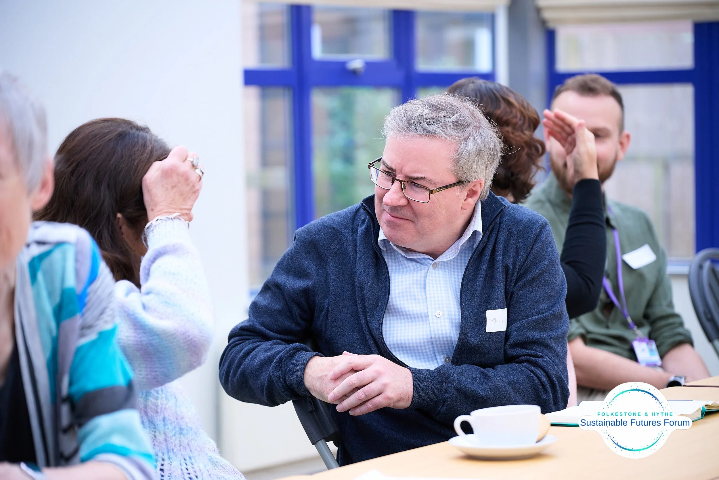 A man in glasses and a blue sweater sitting at a table, grimacing while talking to a woman with dark hair who is holding her head. Other people are visible in the background during a forum event.