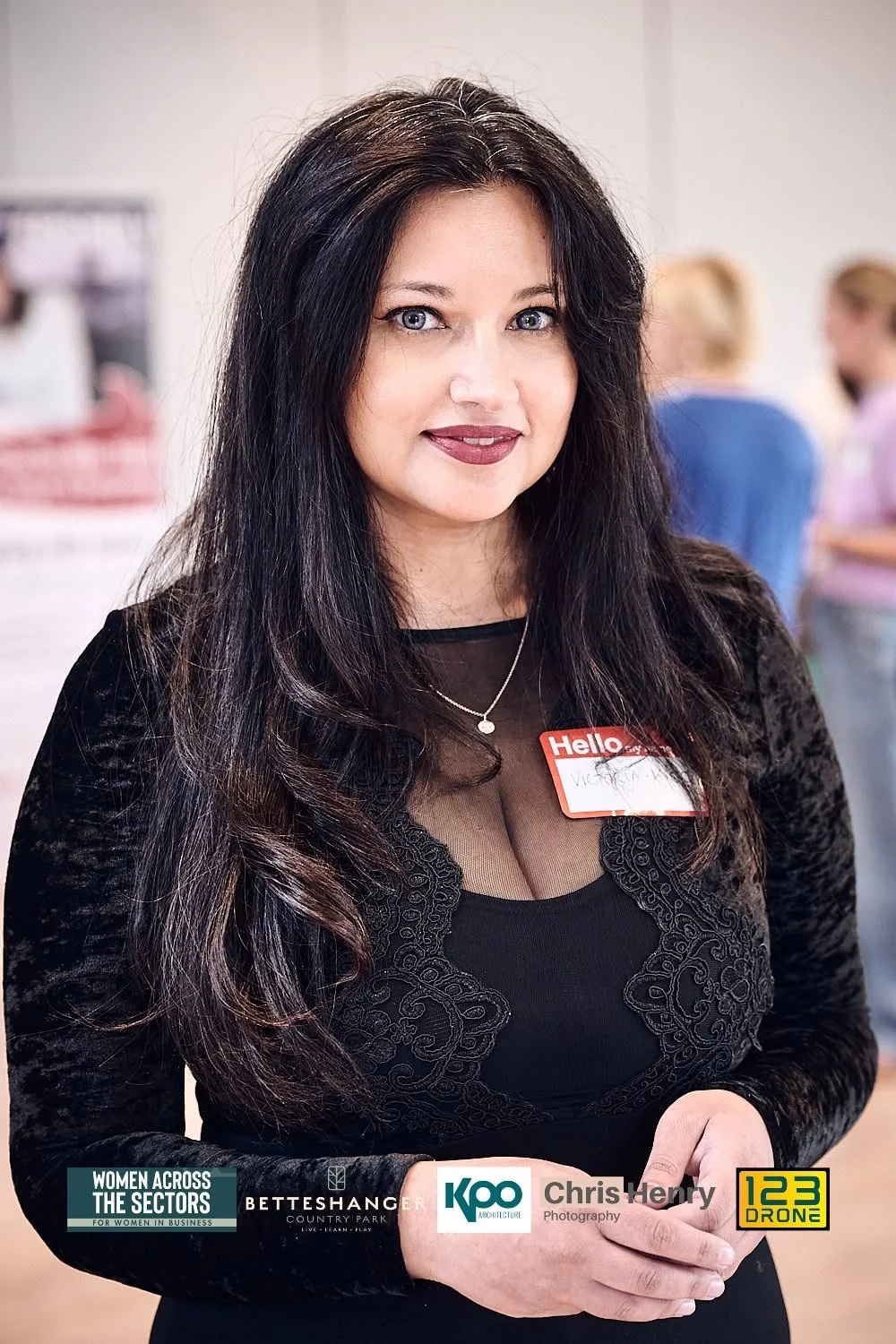 Woman with long dark hair wearing a black dress with lace detailing, standing at an event with a name tag that says 'Hello' and smiling at the camera, with a blurred background of other people.