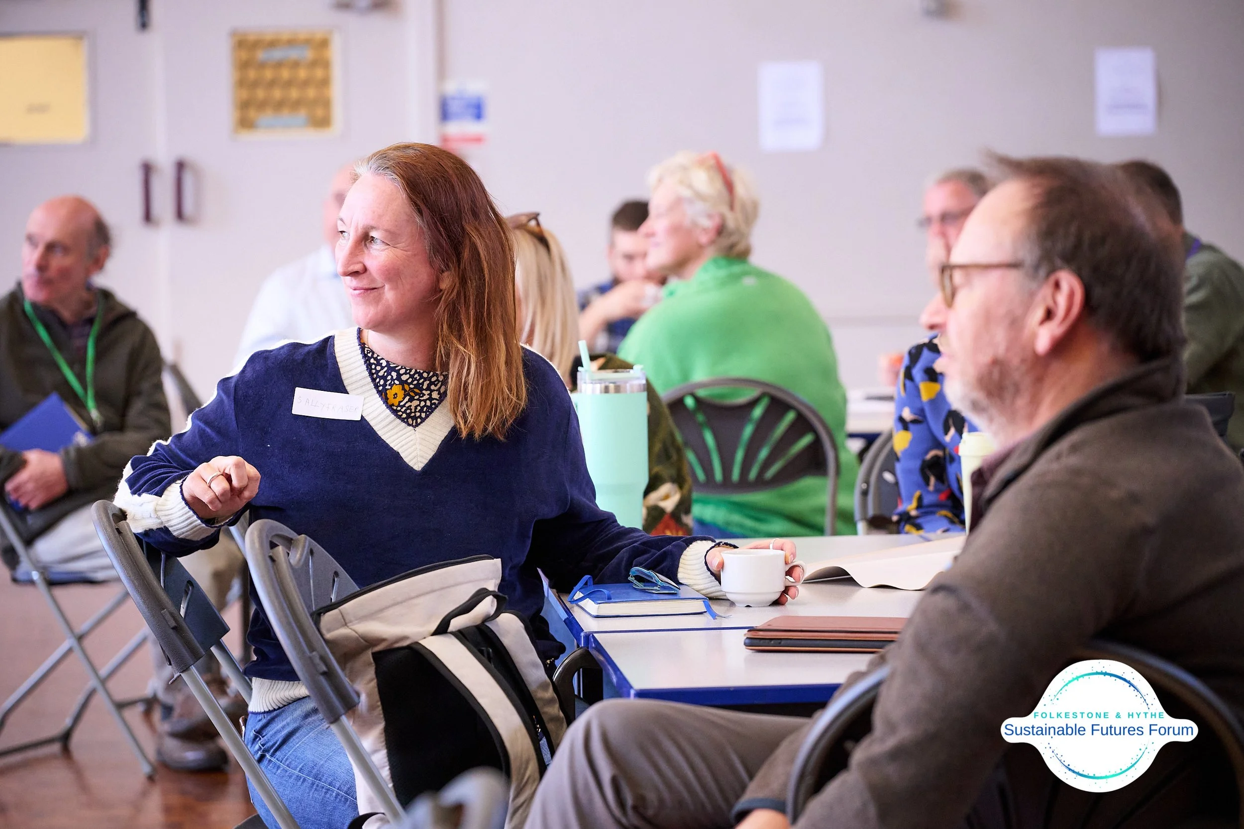 People attending a community or conference event, sitting at tables in a meeting room, engaged in conversations. A woman with red hair and a woman with glasses are prominent in the foreground, with notes and coffee on the table.