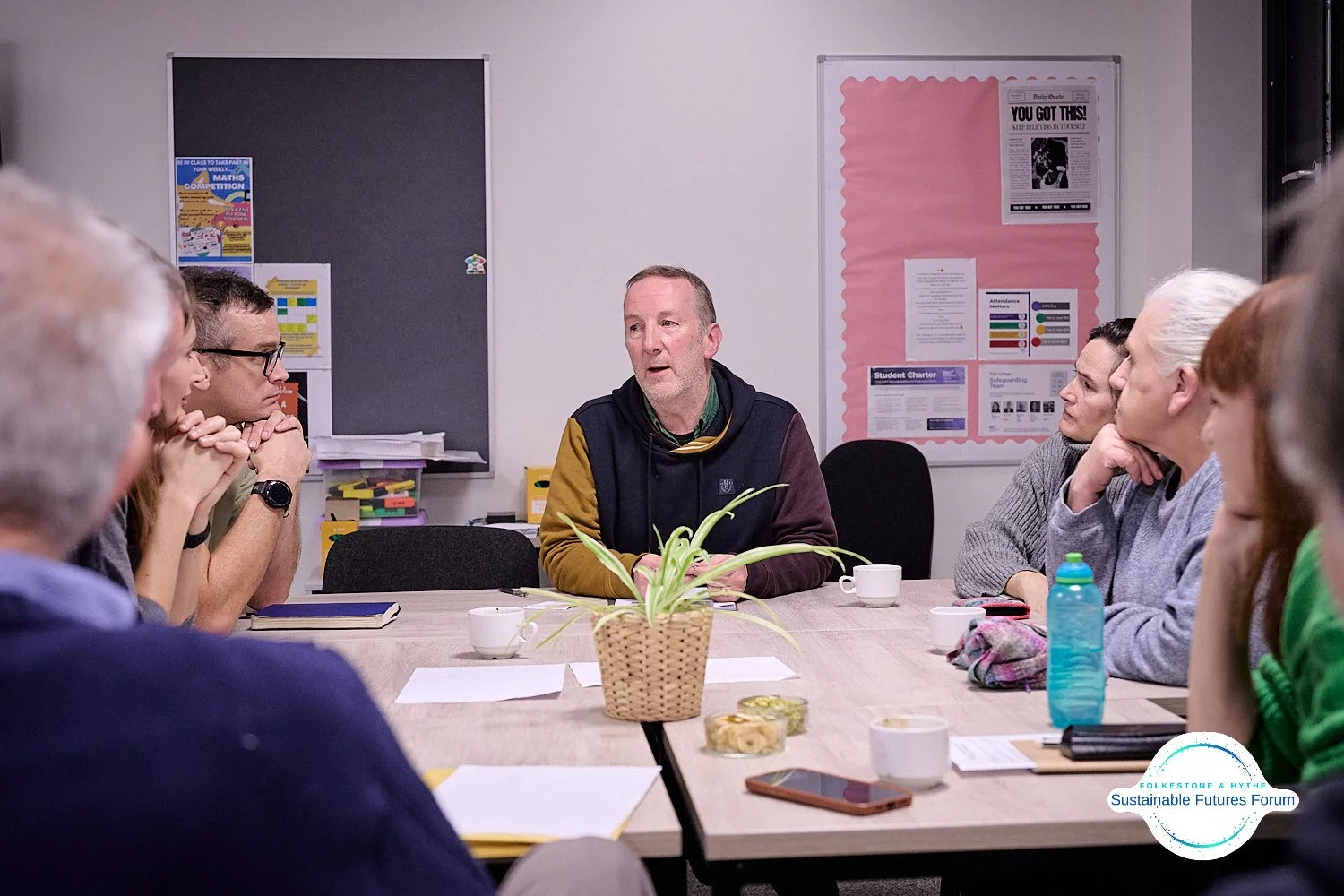 Group of people sitting around a conference table in a meeting room, listening to a speaker. The table has papers, cups, a water bottle, and snacks. A potted plant is in the center of the table. Bulletin boards are on the walls in the background.