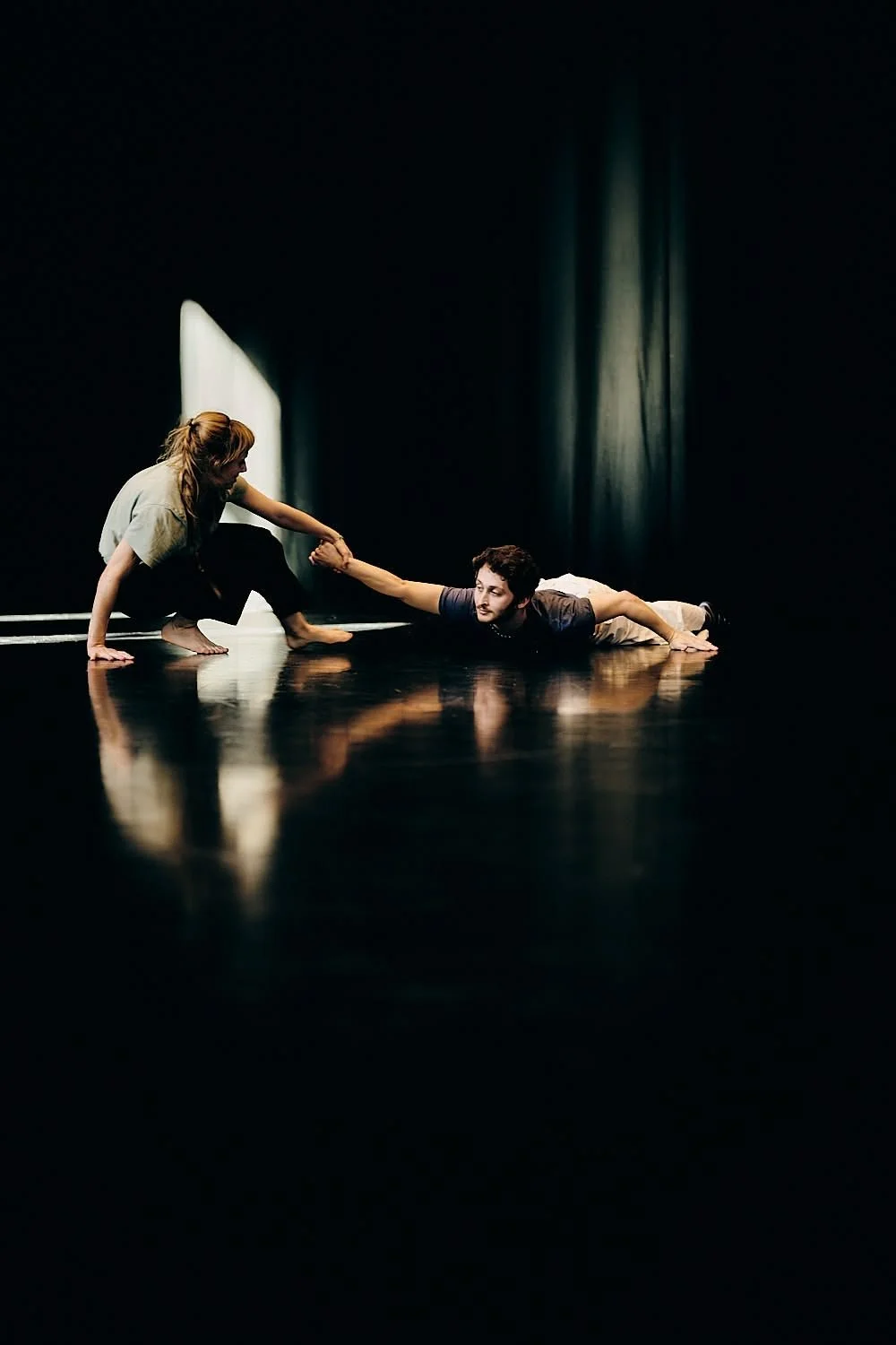 Two people performing a dance move on a stage with black curtains, one extending a hand to the other, who is lying on the floor.