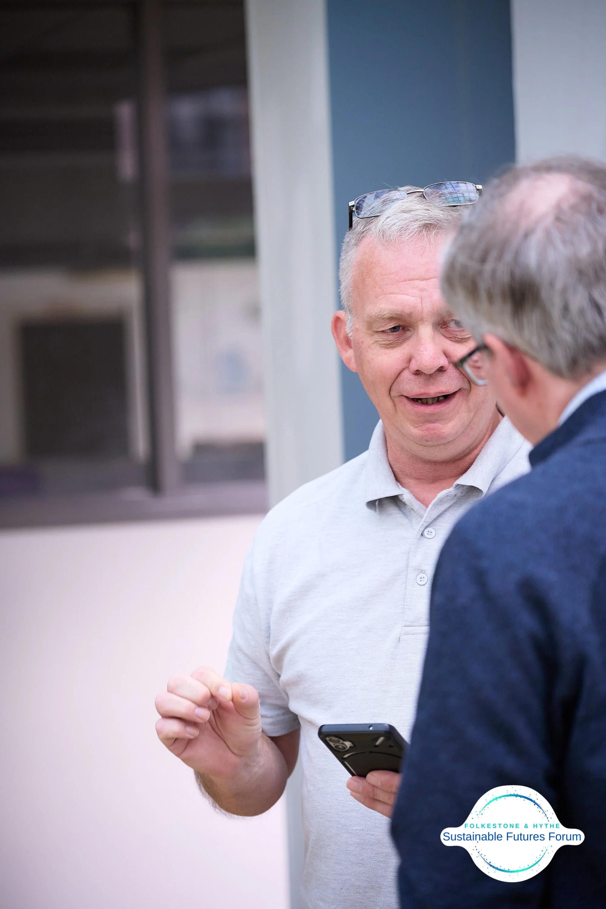 Two middle-aged men are having a conversation outdoors. One man, wearing a light gray polo shirt and glasses on his head, is holding a smartphone and gesturing with his hand. The other man, wearing a dark jacket and glasses, has gray hair. There is a