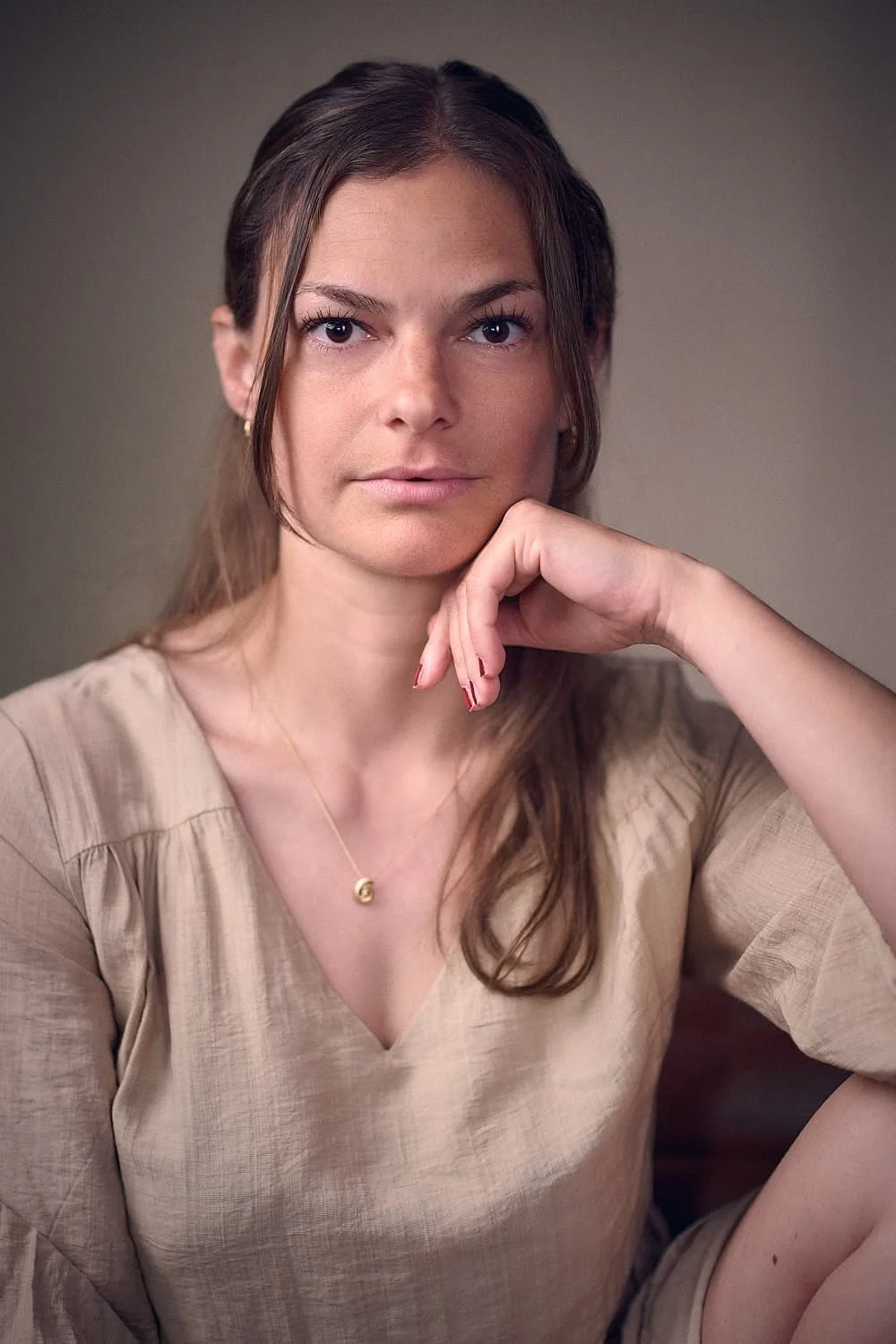 Portrait of a woman with brown hair, wearing a beige top and a delicate gold necklace, resting her chin on her hand, looking directly at the camera.
