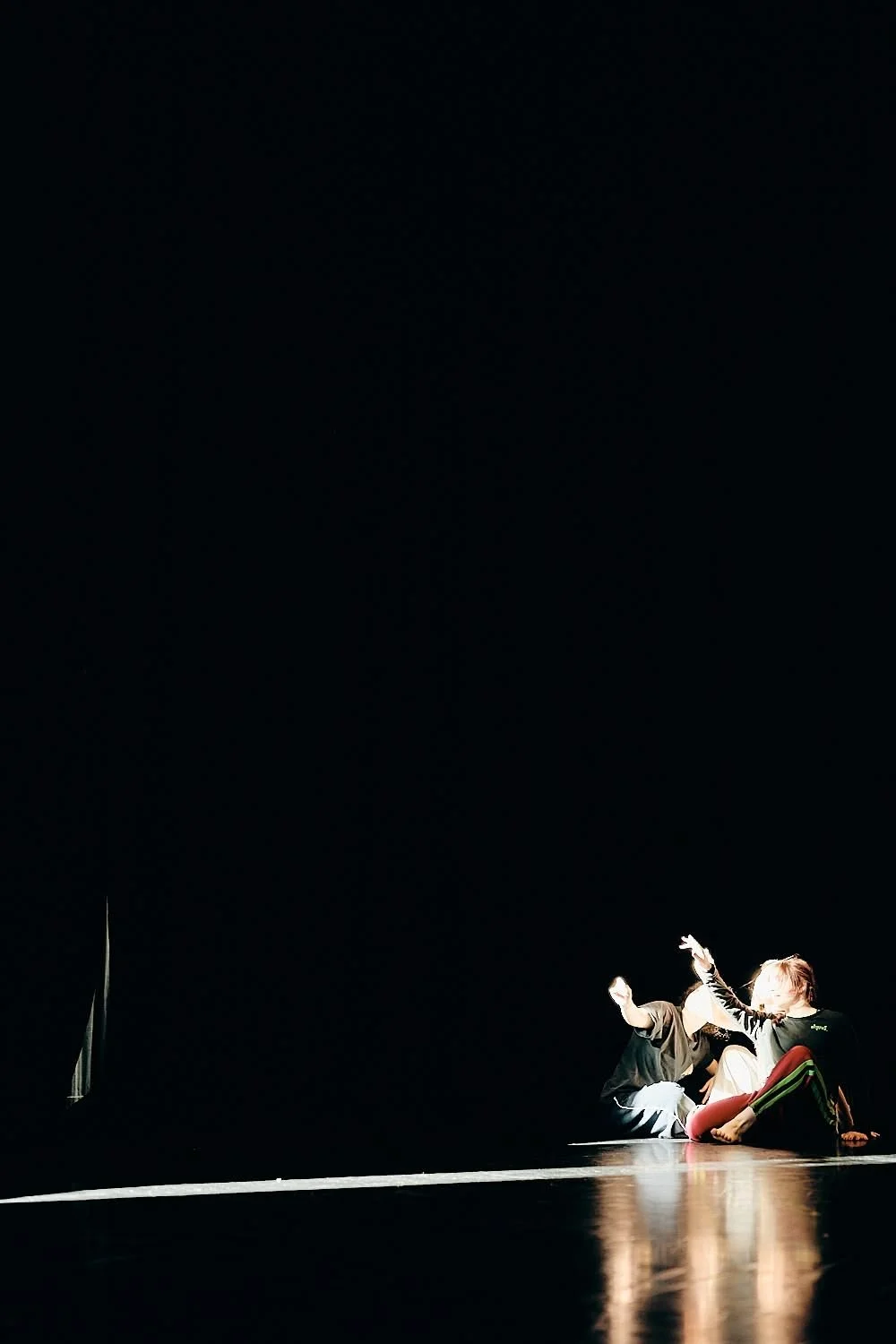 Two performers on stage, sitting in a dark setting, with one person reaching out towards the other, illuminated by a spotlight.