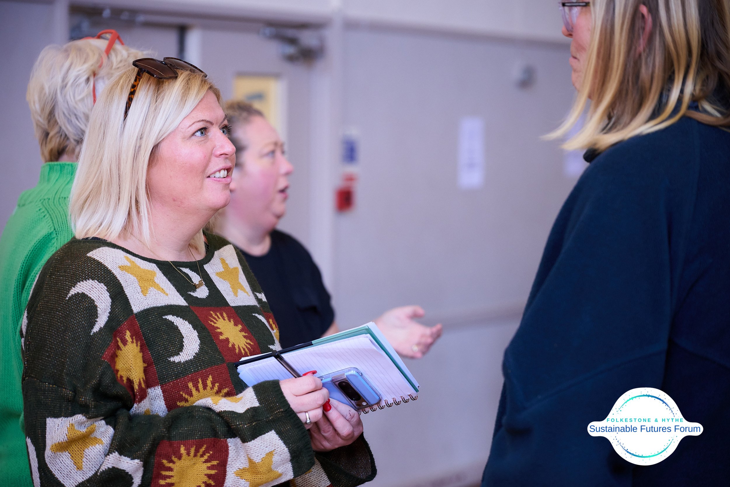 A group of women, including one wearing a patterned sweater with suns, moons, and stars, are engaged in conversation at a forum or conference, with the logo 'Folkestone & Hythe Sustainable Futures Forum' visible in the corner.