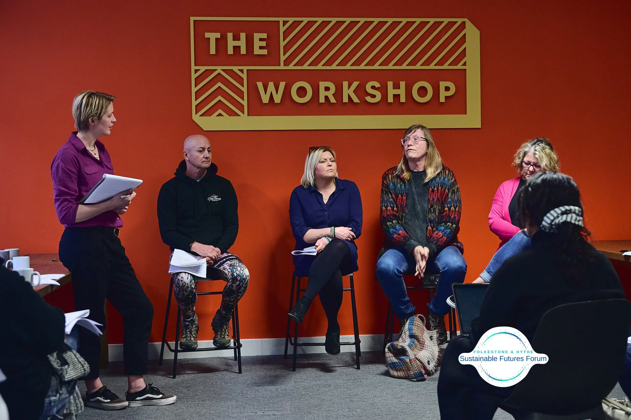Panel discussion at The Workshop with five women seated and one standing, against an orange wall with a large sign that says "The Workshop." There are people in the audience, some with notebooks and laptops.