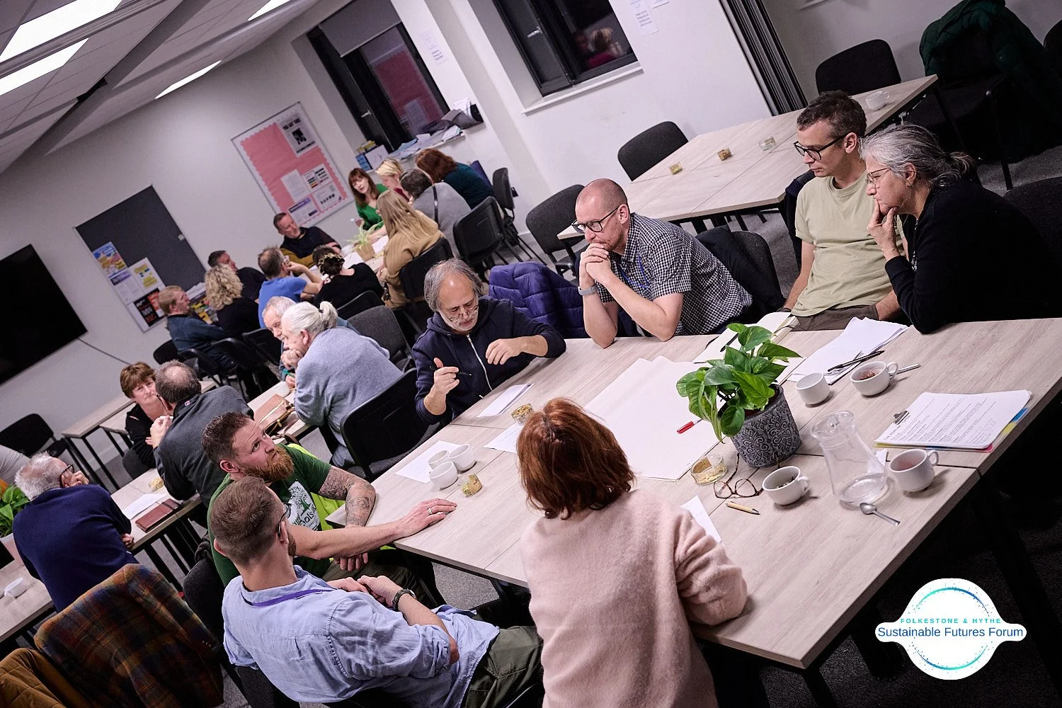 People gathered at tables in a meeting room, engaged in discussions, with papers, notebooks, cups, and a potted plant on the table in the foreground.