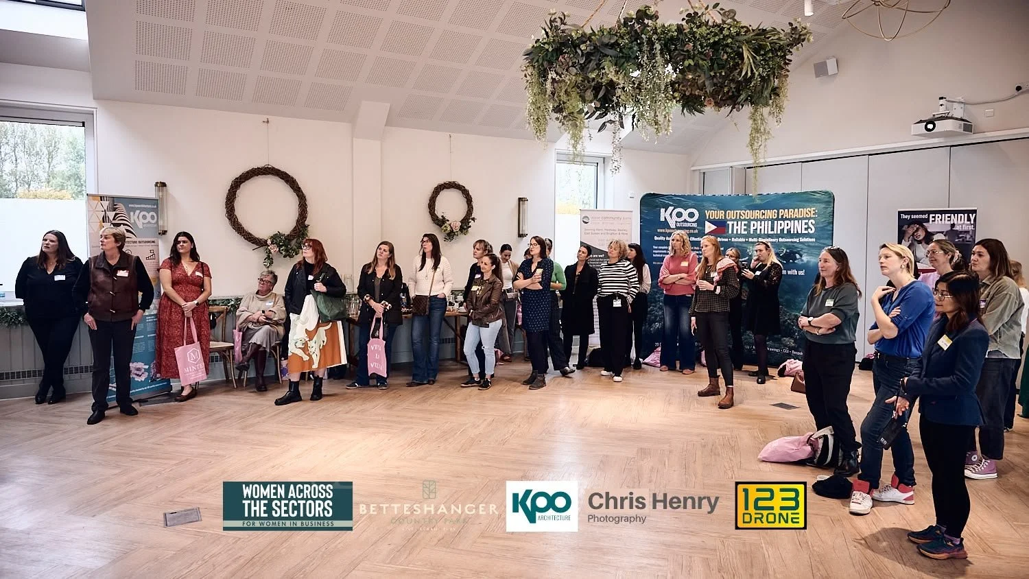 Group of women standing in a semi-circle at an indoor event. Some are listening and watching, others are taking photos or notes. The room is decorated with floral wreaths and hanging greenery, with banners and signs on the walls promoting women in bu