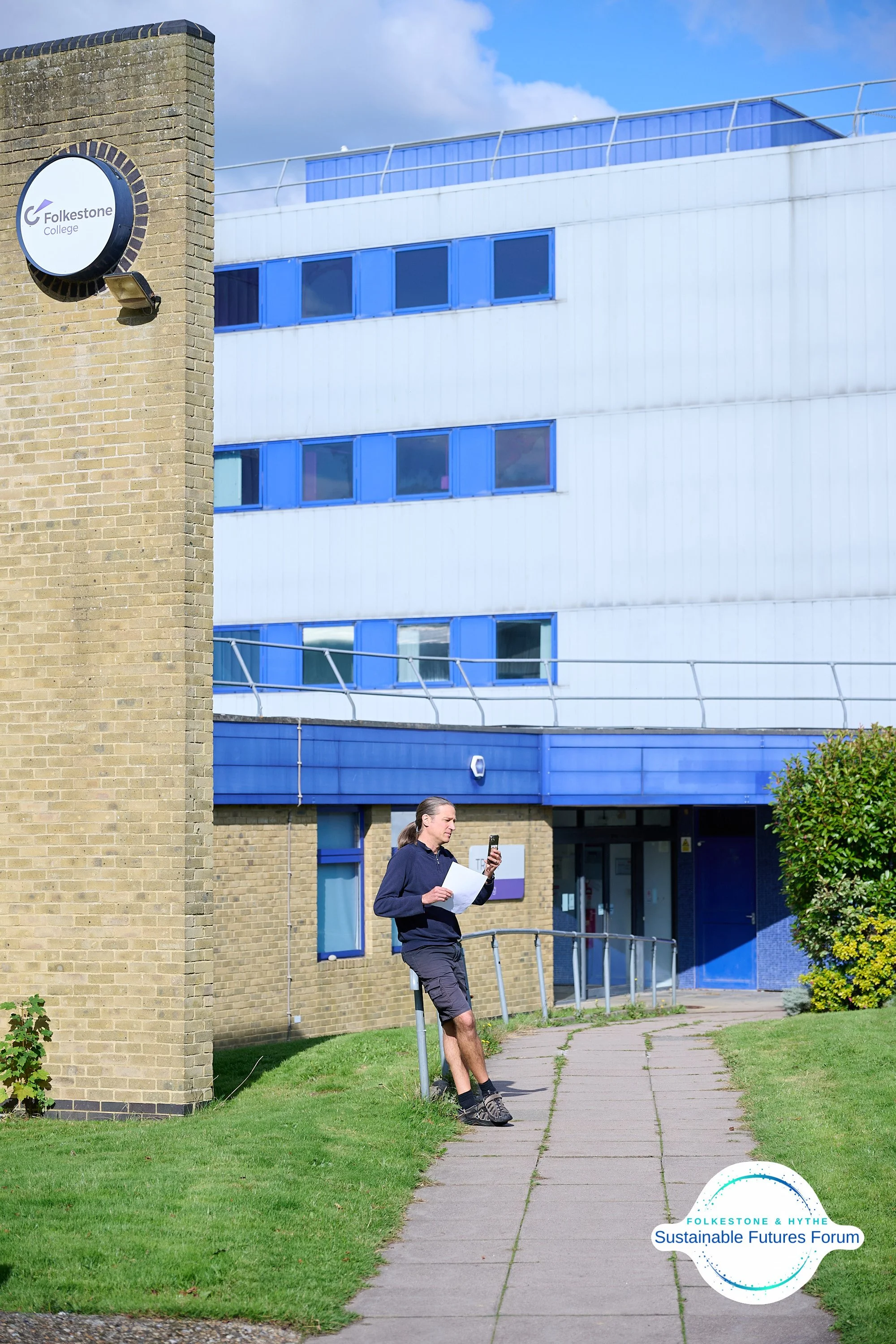 A person leaning against a railing outside a modern building with blue windows, holding a phone and papers. A sign on the building reads 'Folkestone College,' and there is a logo in the bottom right corner that says 'Folkestone & Hythe Sustainable Fu