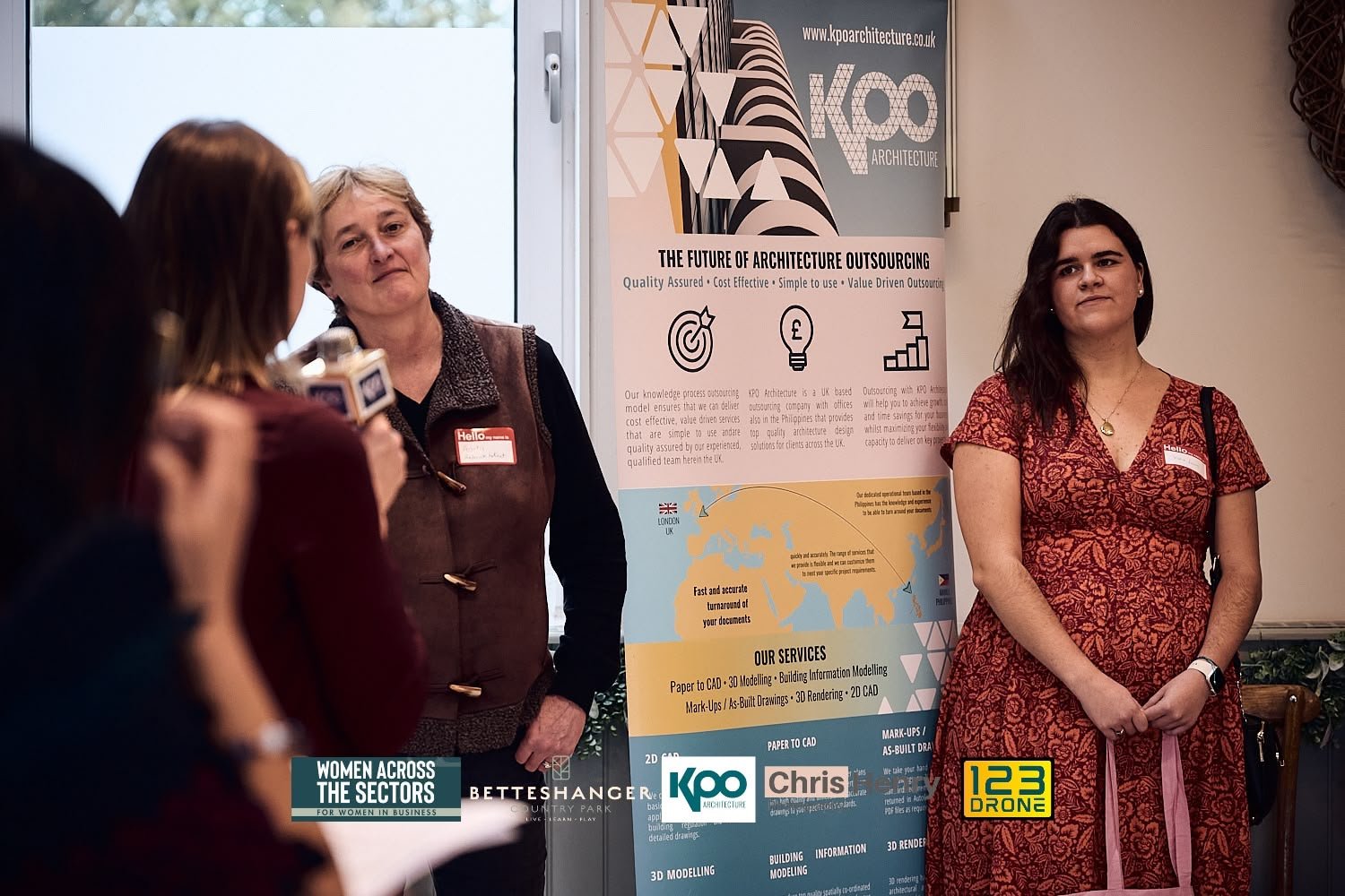 Women at a presentation or exhibition stand about architecture outsourcing, with a large informational poster behind them, and a woman speaking into a microphone. One woman is wearing a red patterned dress, another is in a dark vest, and a third woma