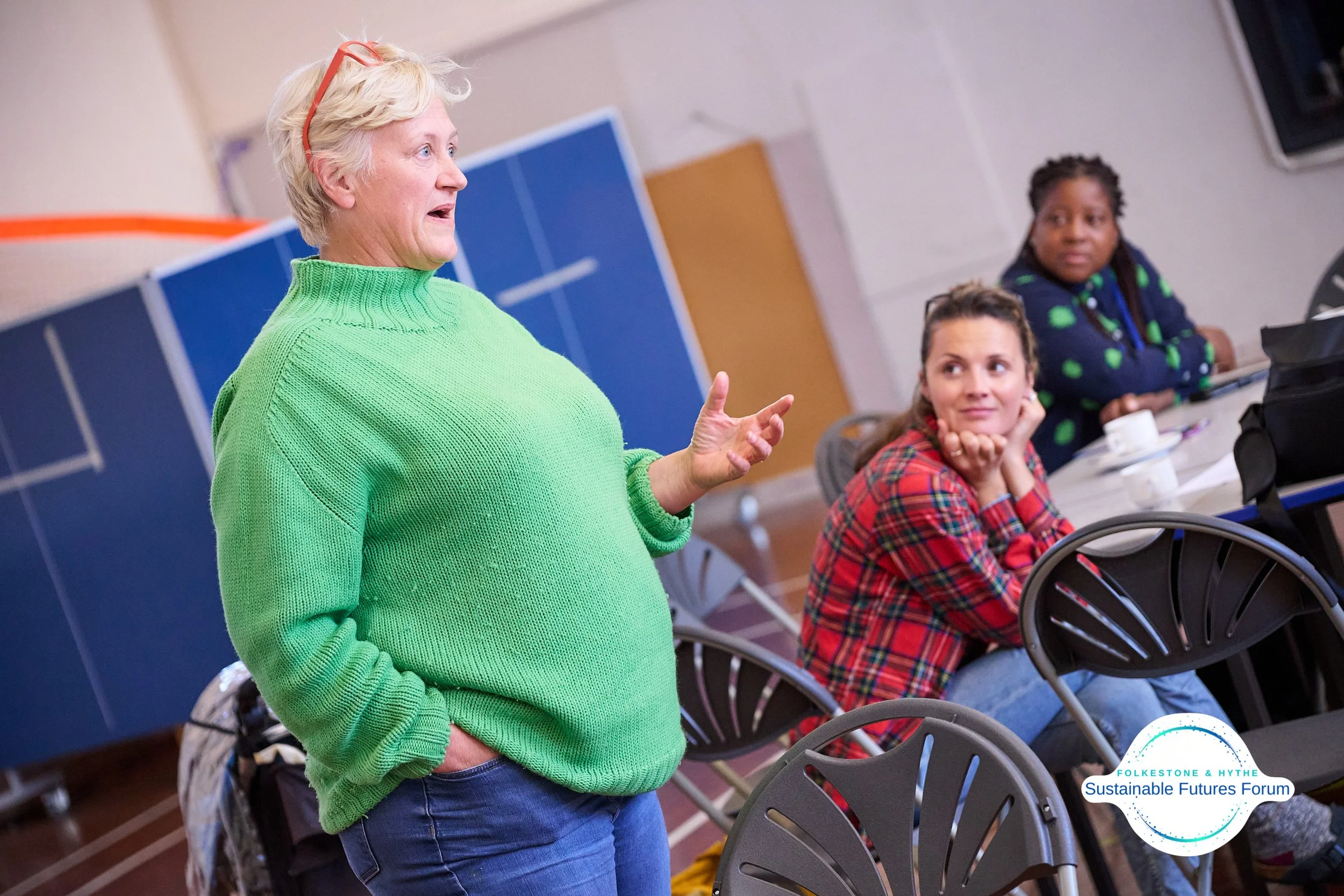 A woman with short blonde hair wearing a bright green sweater speaking to a small group of women seated at a table in a conference room. One of the women in a red plaid shirt is looking at her attentively, while another woman with braided hair and a 