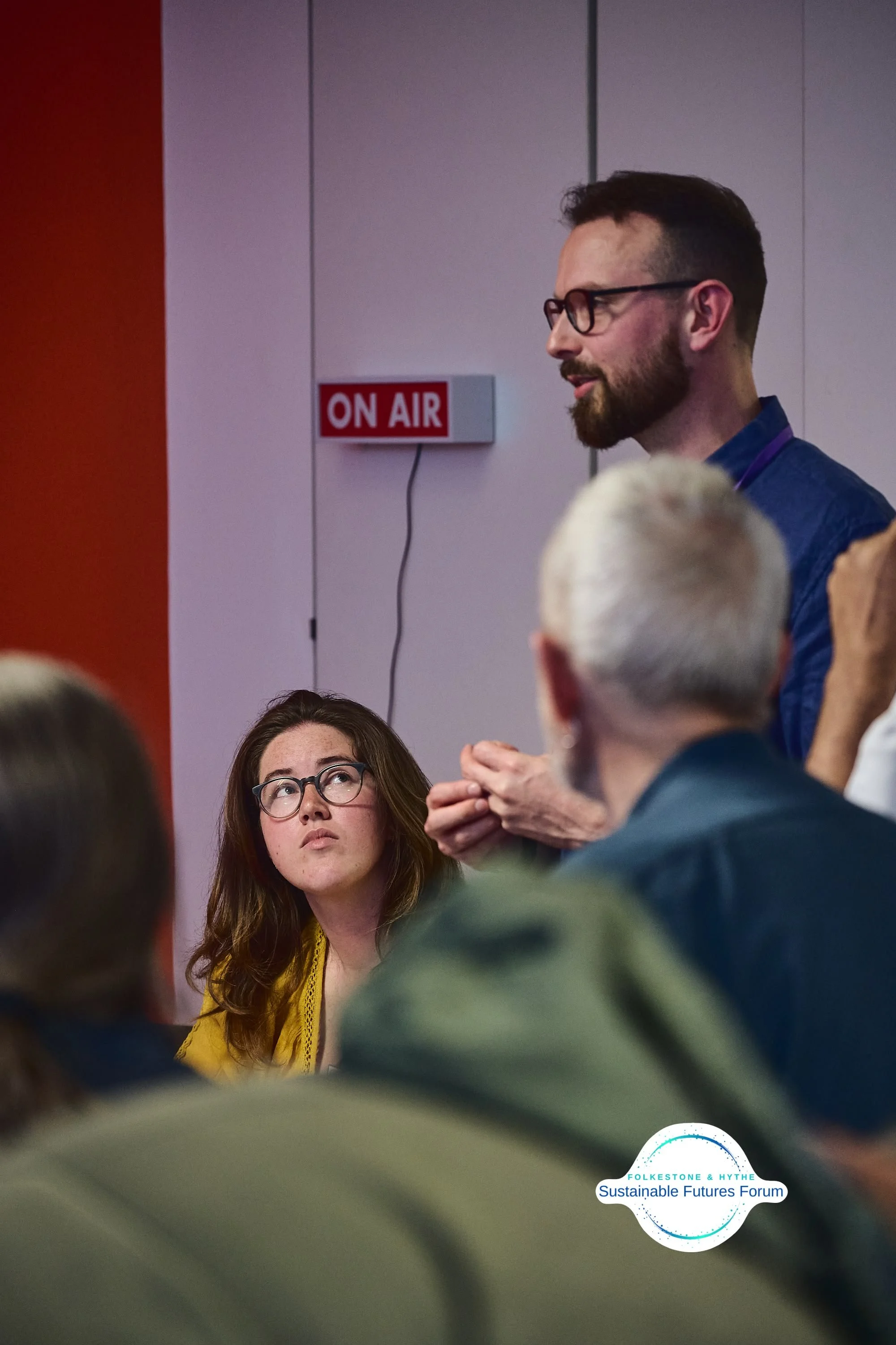 A man with glasses and a beard speaking at a podium during a forum, with a woman wearing glasses looking on. The forum is part of the Folkestone & Hythe Sustainable Futures Forum, as indicated by the logo. An 'ON AIR' sign is visible on the wall behi