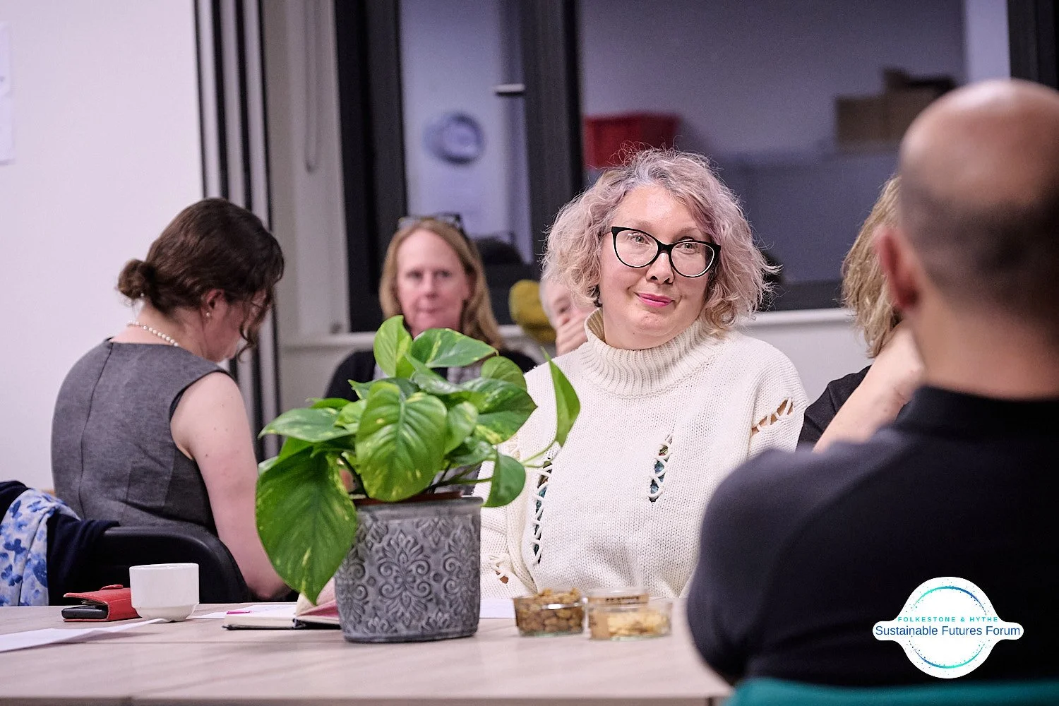 A group of people sitting around a table engaging in conversation, with a woman with curly blonde hair and glasses as the focal point, a potted plant on the table, and the logo of the Folkestone & Hythe Sustainable Futures Forum in the bottom right c