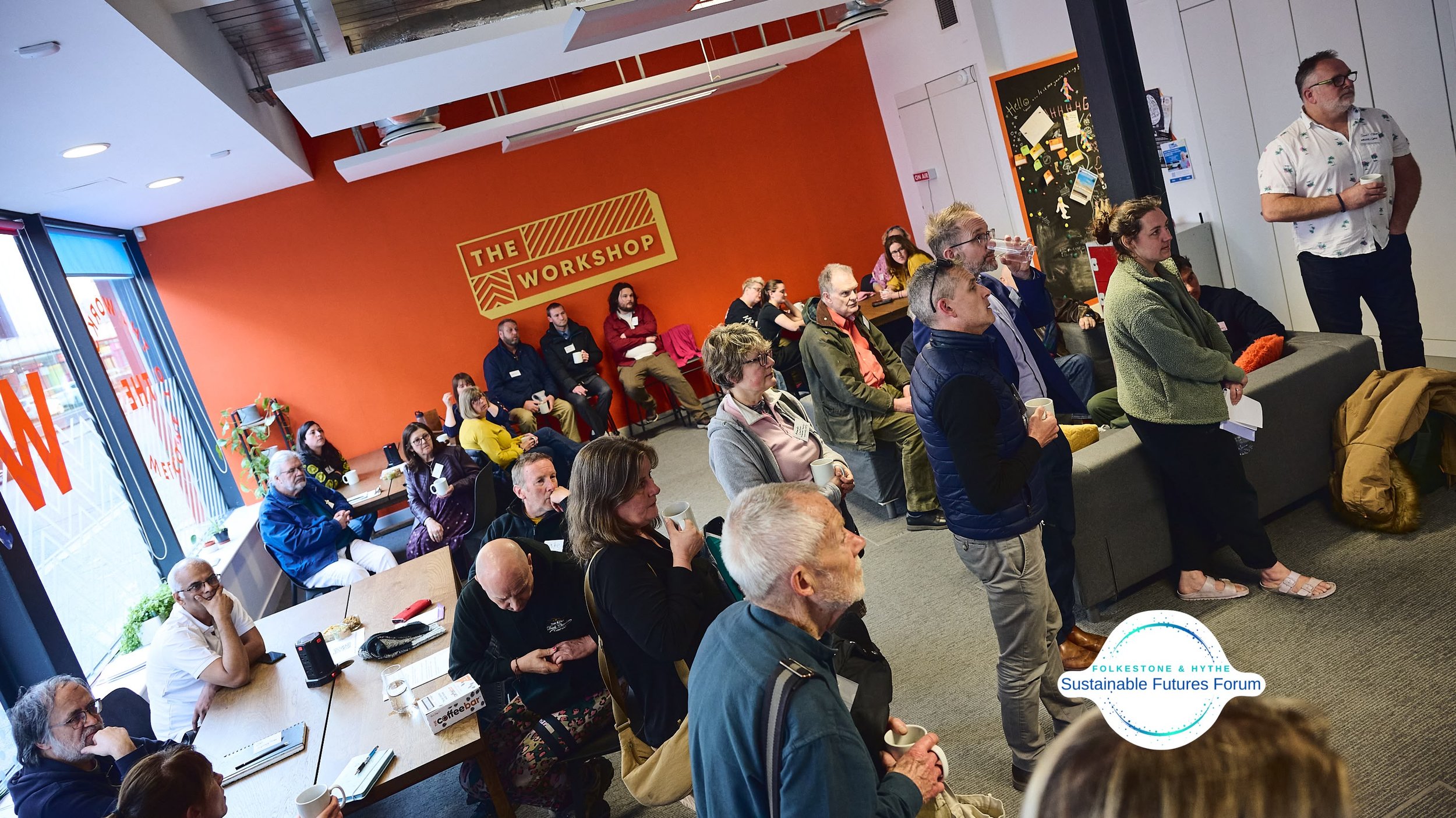 A group of people attending the Folkestone & Hythe Sustainable Futures Forum in a bright room with an orange wall and a sign that says 'The Workshop.' Some people are sitting, others are standing, and one person is speaking. There are windows on the 