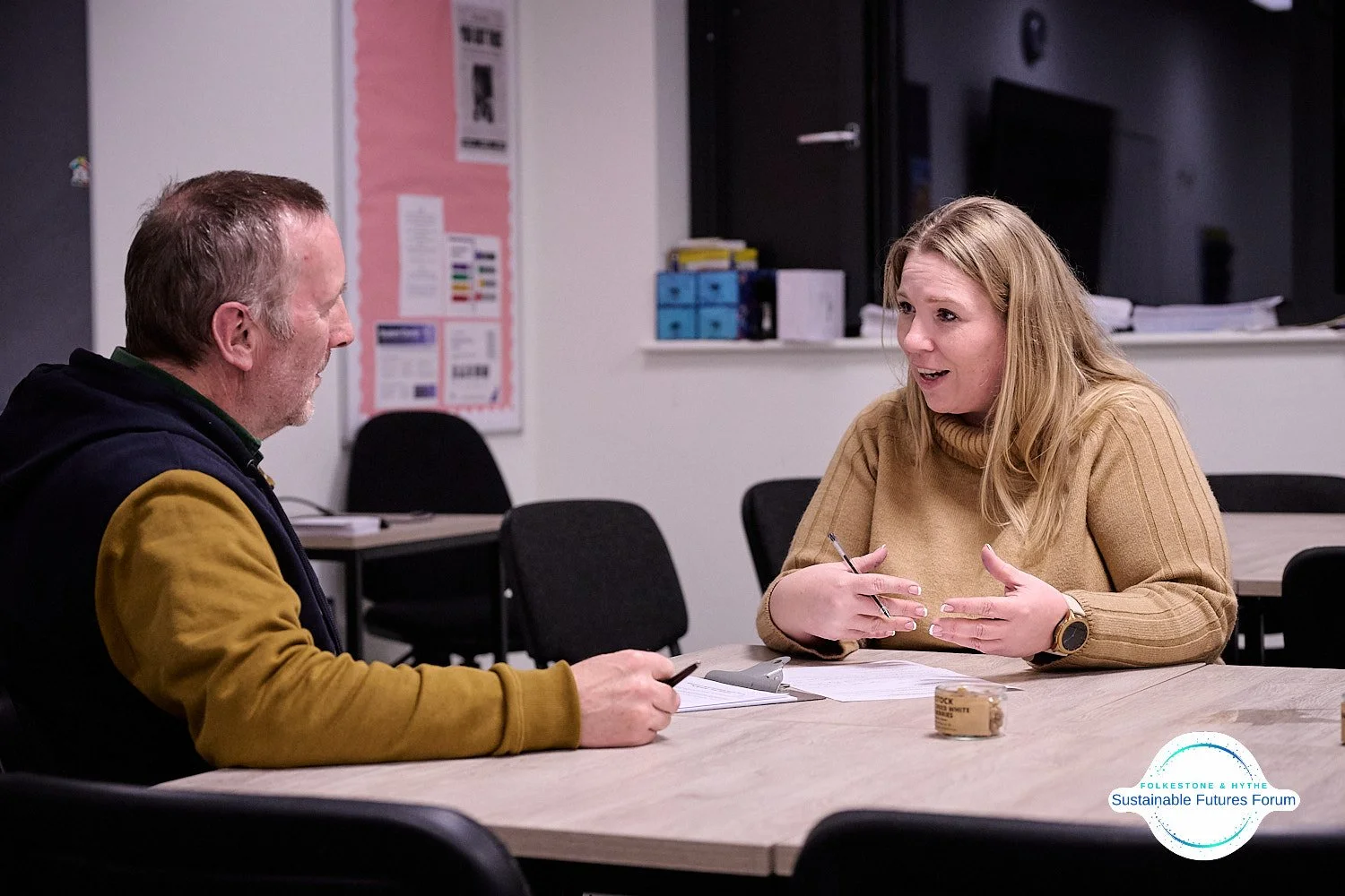 Two people having a conversation at a table in a classroom or office, with papers and small objects on the table.