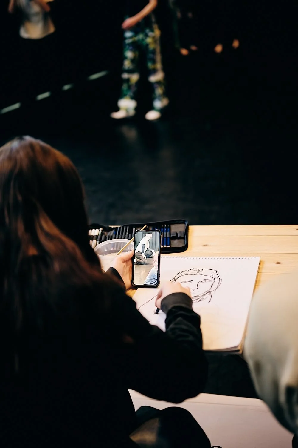 A woman sitting at a desk sketching a portrait of a person on a notepad, while taking a photo of a person in the background with her phone. The desk has drawing supplies and the background is dark with a person standing across.