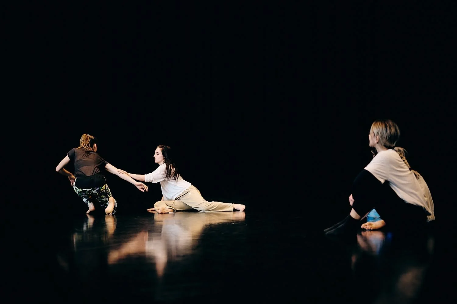 Three women on a dark stage, with two of them sitting on the floor and one kneeling, engaged in a dance or theatrical performance.