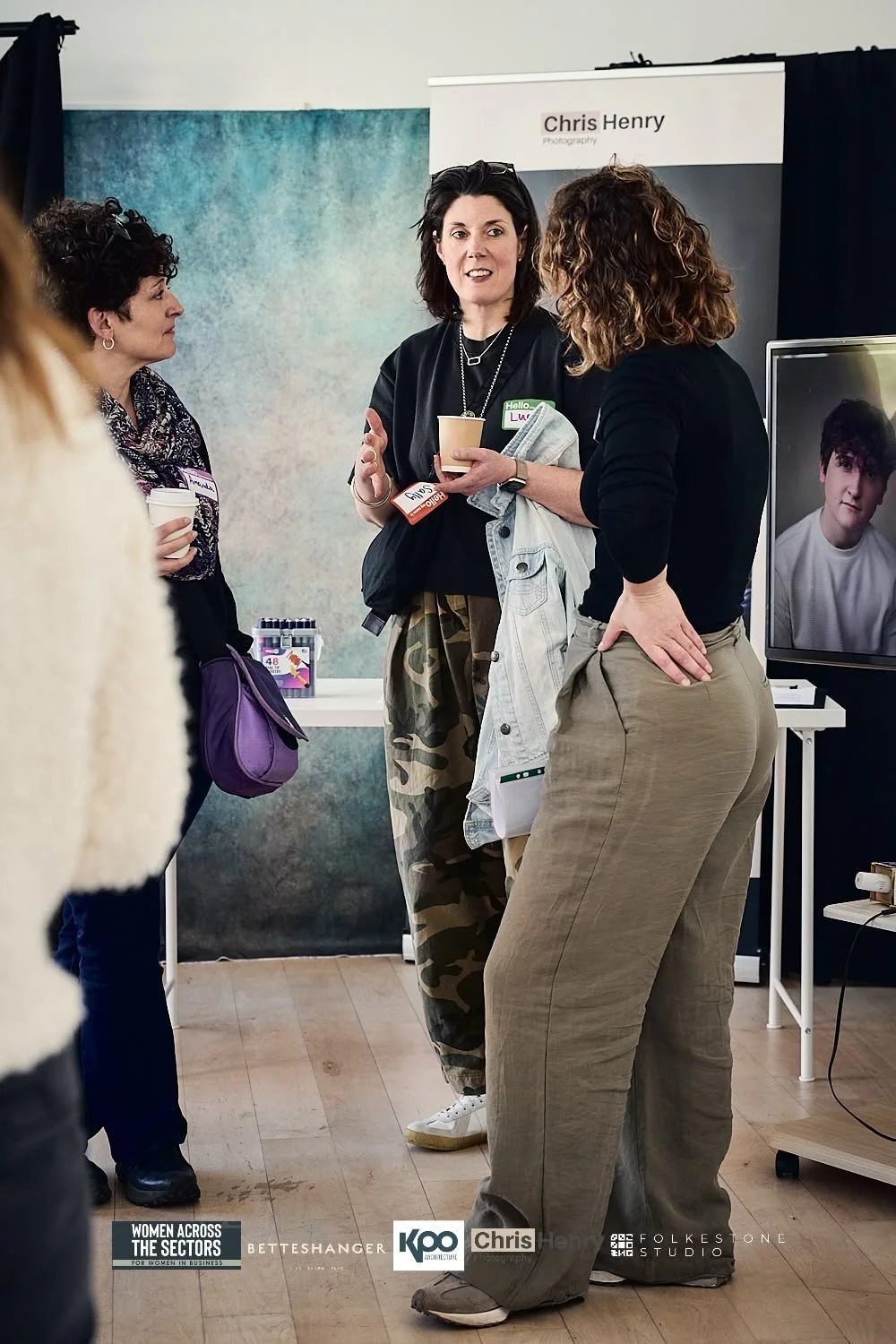 Three women engaging in a conversation at an event, with a banner for Chris Henry photography in the background and a TV screen displaying a portrait of a young man.