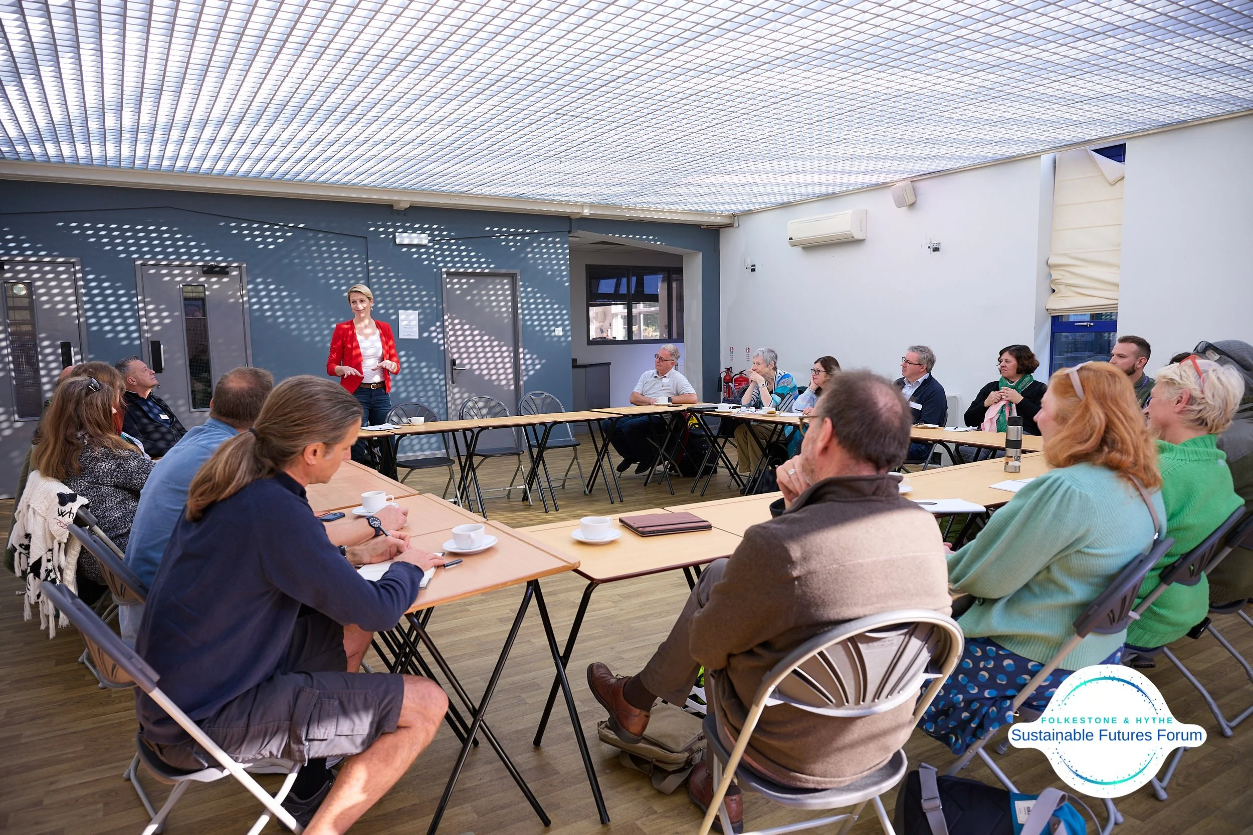 A woman in a red blazer giving a presentation to a group of people seated around a U-shaped table in a well-lit room at the Folketone & Hythe Sustainable Futures Forum.