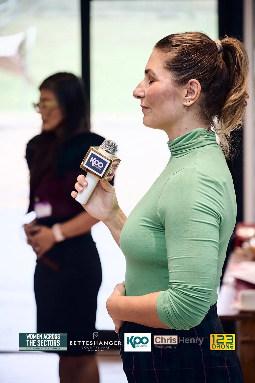 A woman in a green top holding a microphone with the logo 'KOO' during a presentation or event, with another woman blurred in the background.