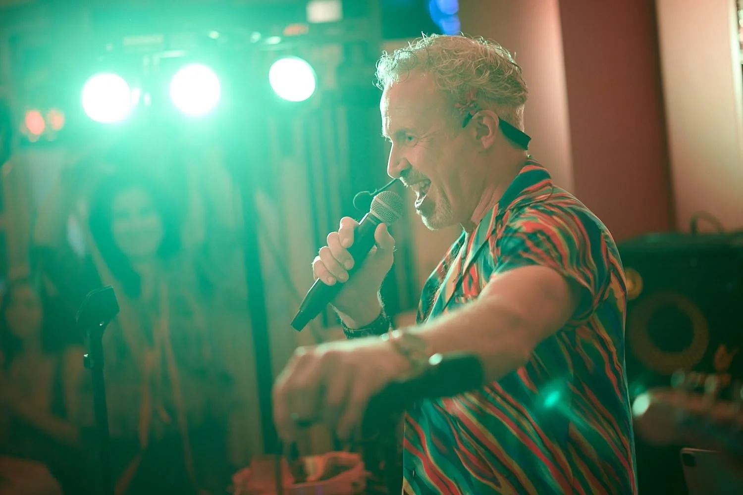 A man with curly hair wearing a colorful shirt, holding a microphone, singing or speaking at a lively event with bright stage lights and people in the background.