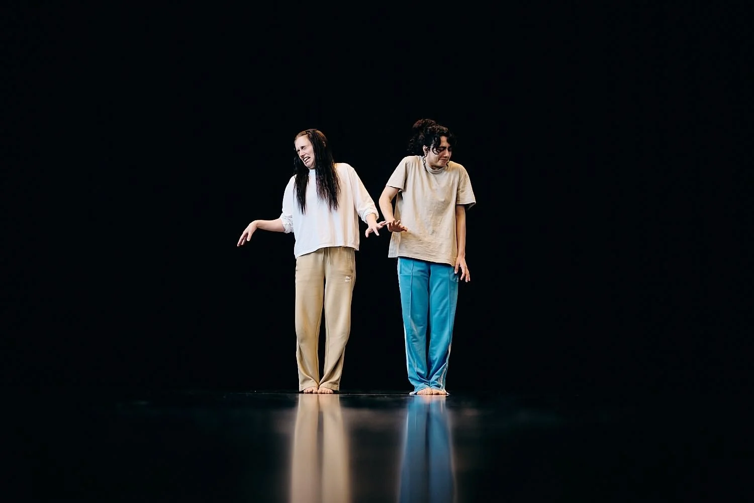 Two women standing barefoot on a stage with a dark background, engaged in a theatrical performance, holding hands and making expressive gestures.