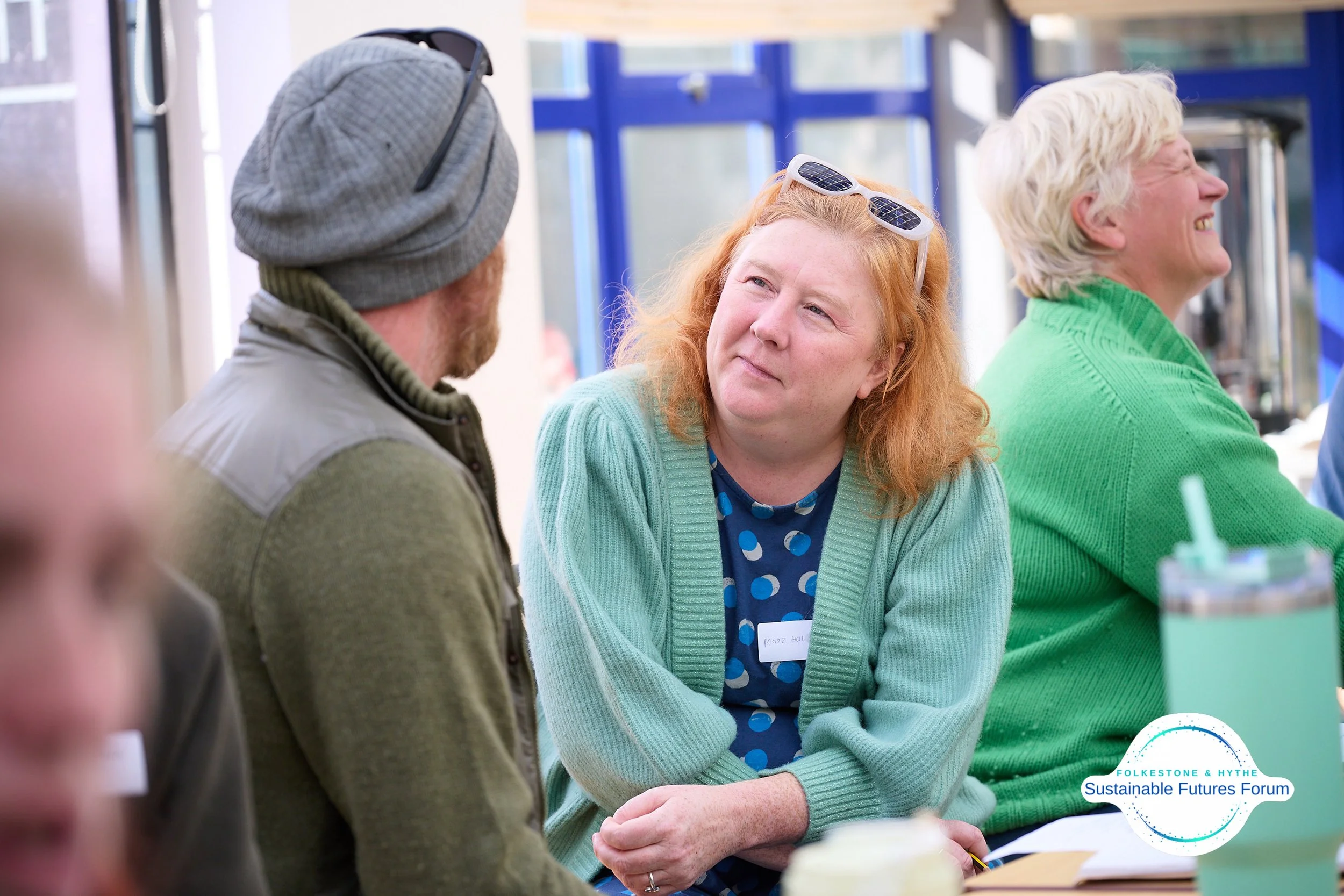 Three adults sitting at a table during a forum, with a woman in a blue polka-dot shirt and green cardigan talking to a man in a gray beanie and vest, and an older woman in a green sweater smiling.