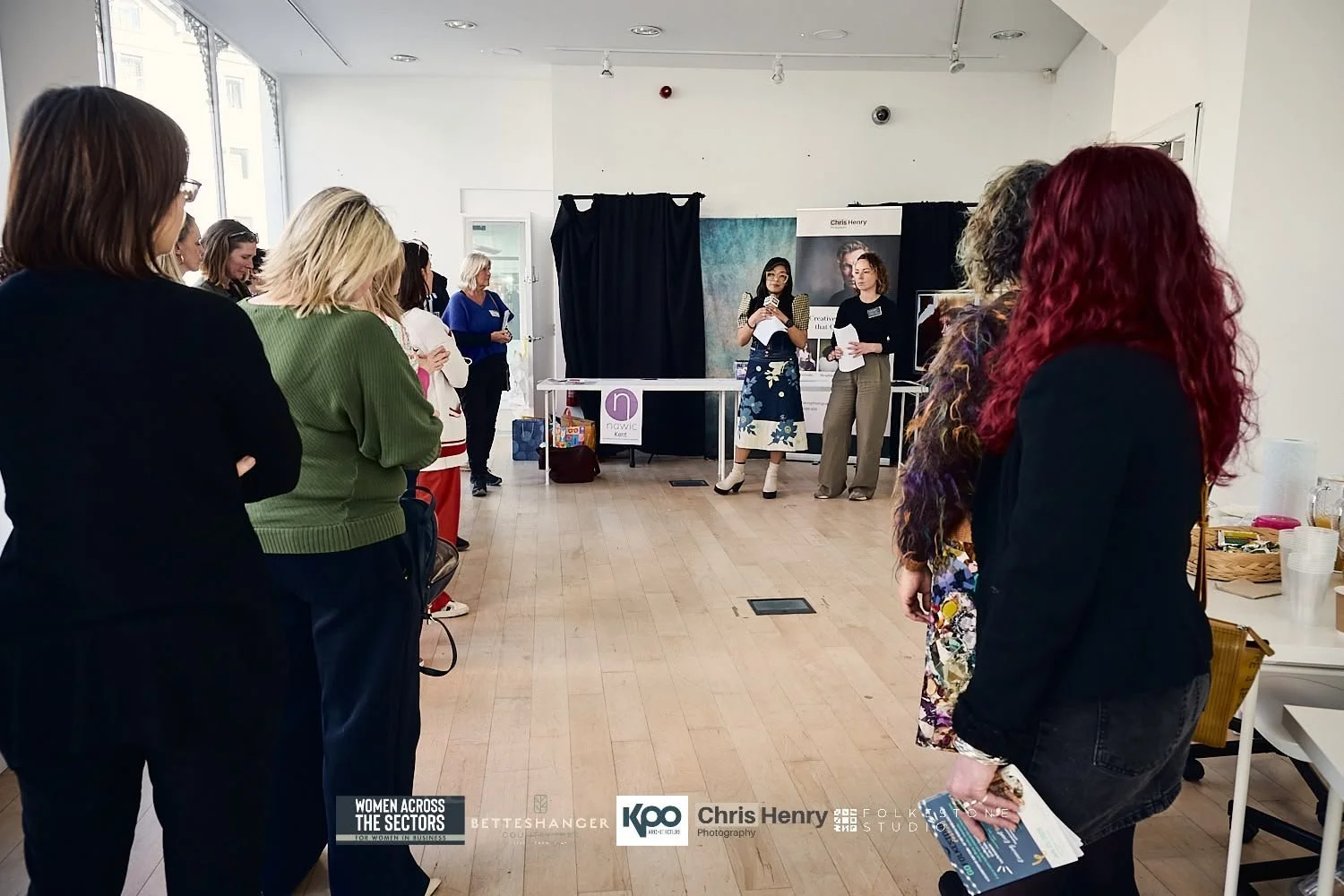 Women attending a presentation or workshop in a spacious, well-lit room with light wooden floors, standing in a semi-circle facing two women speaking at the front, one holding a microphone.