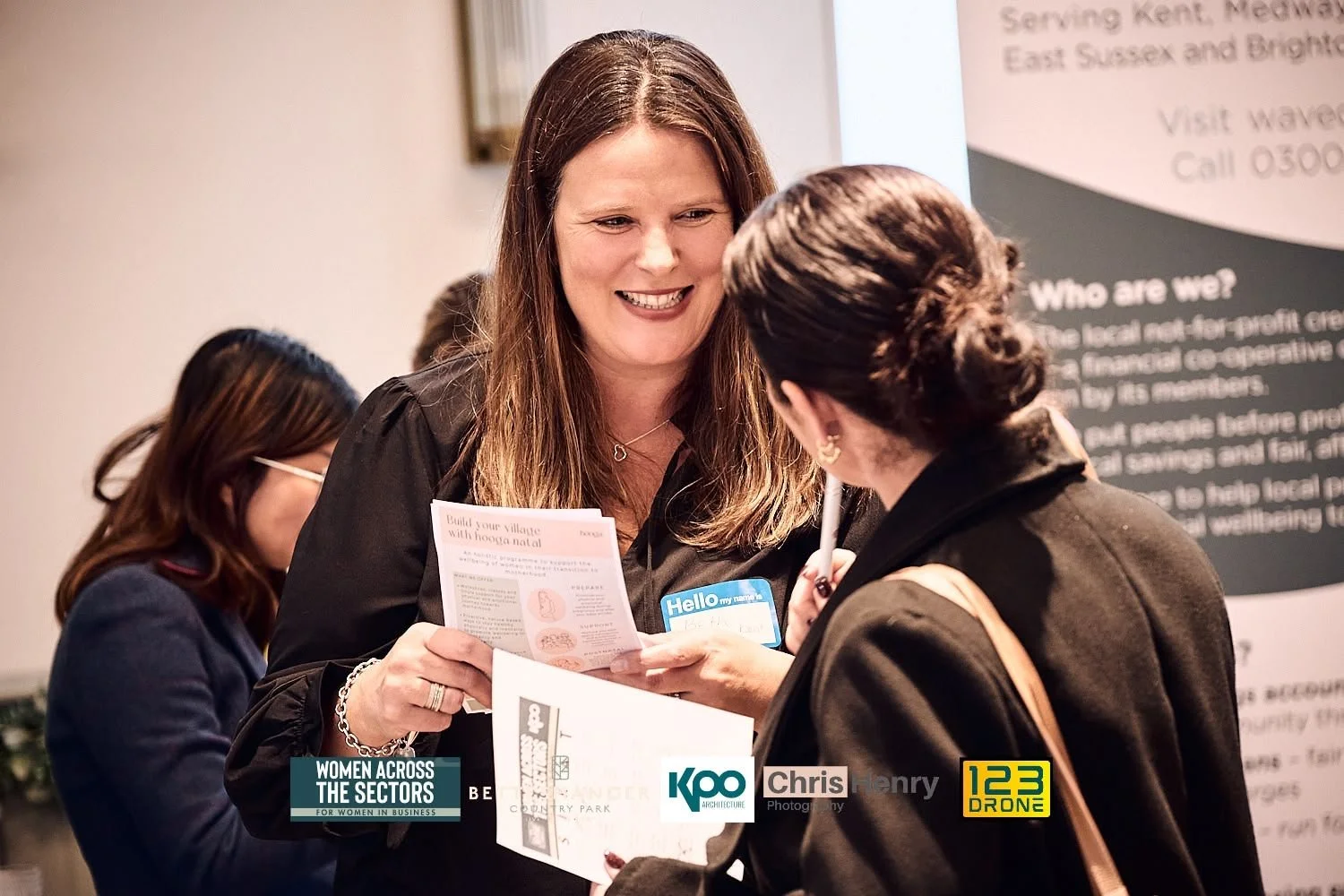 Two women are engaged in conversation at a professional event. The woman facing the camera has long brown hair, is smiling, and is holding a brochure. She is wearing a black jacket and a name tag. The other woman has her back to the camera, her hair 