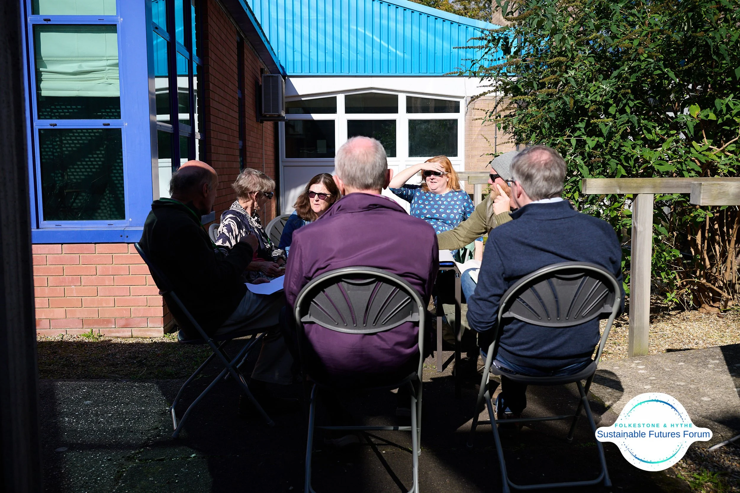 A group of seven people sitting around a table outdoors, engaged in a discussion. The setting appears to be a garden or patio area adjacent to a building with brick and blue siding, with trees and a bush in the background. Sunlight is illuminating th