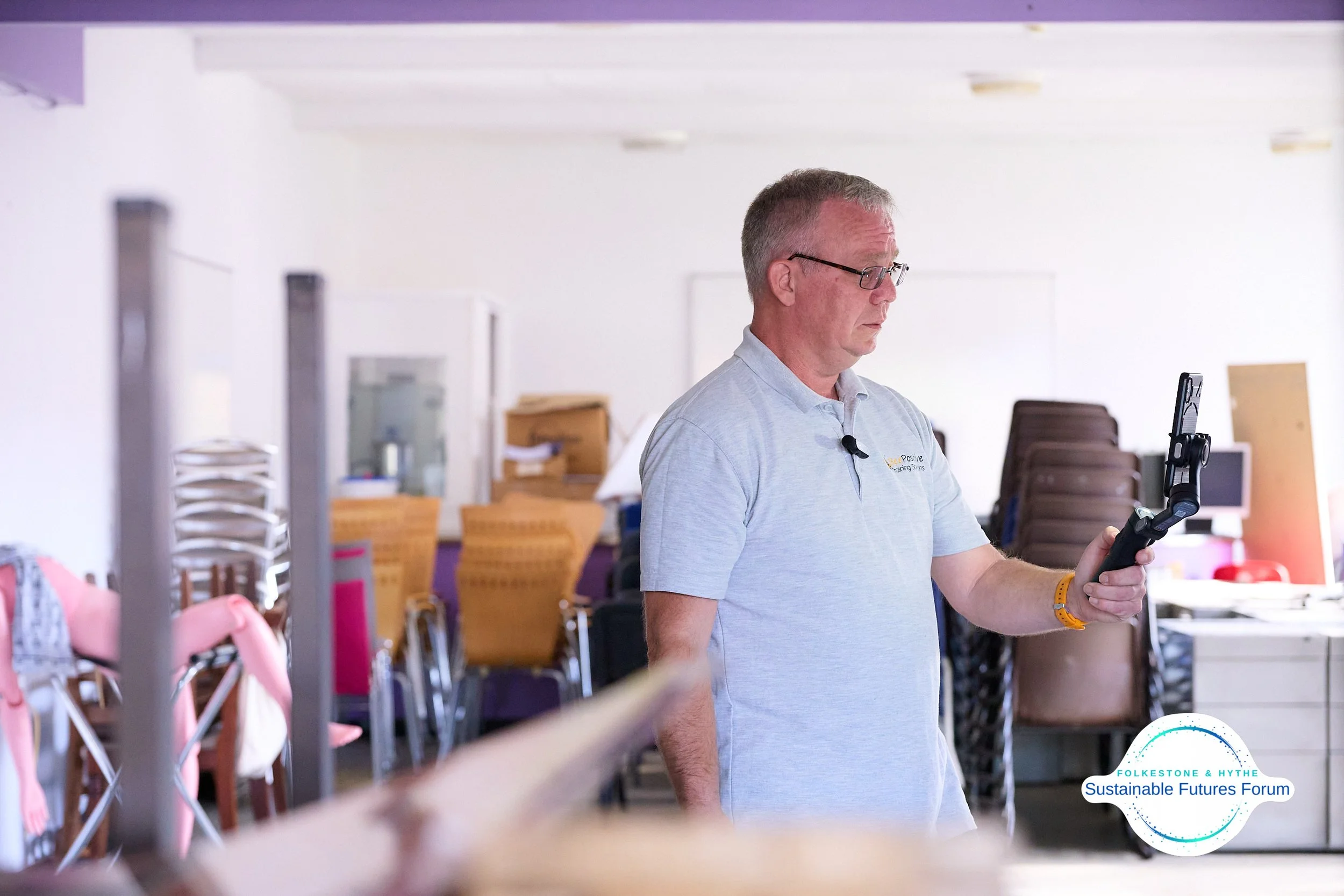 Man wearing glasses and a light gray polo shirt looking at a smartphone in a room with stacked chairs and boxes.