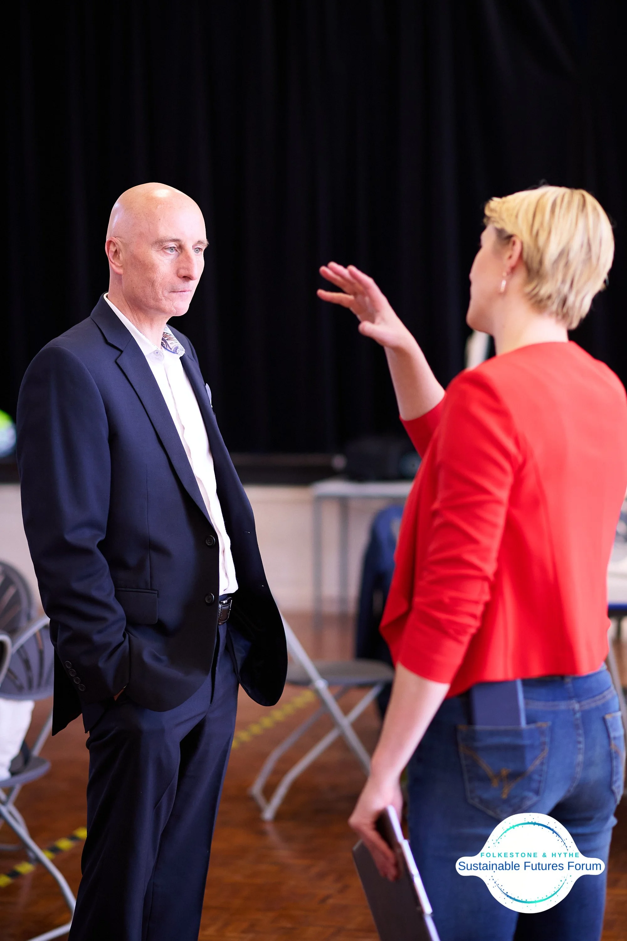 A woman in a red blazer and jeans is talking to a man in a navy suit at an indoor event with chairs in the background. There is a logo for the Folke Stone & Hythe Sustainable Futures Forum.