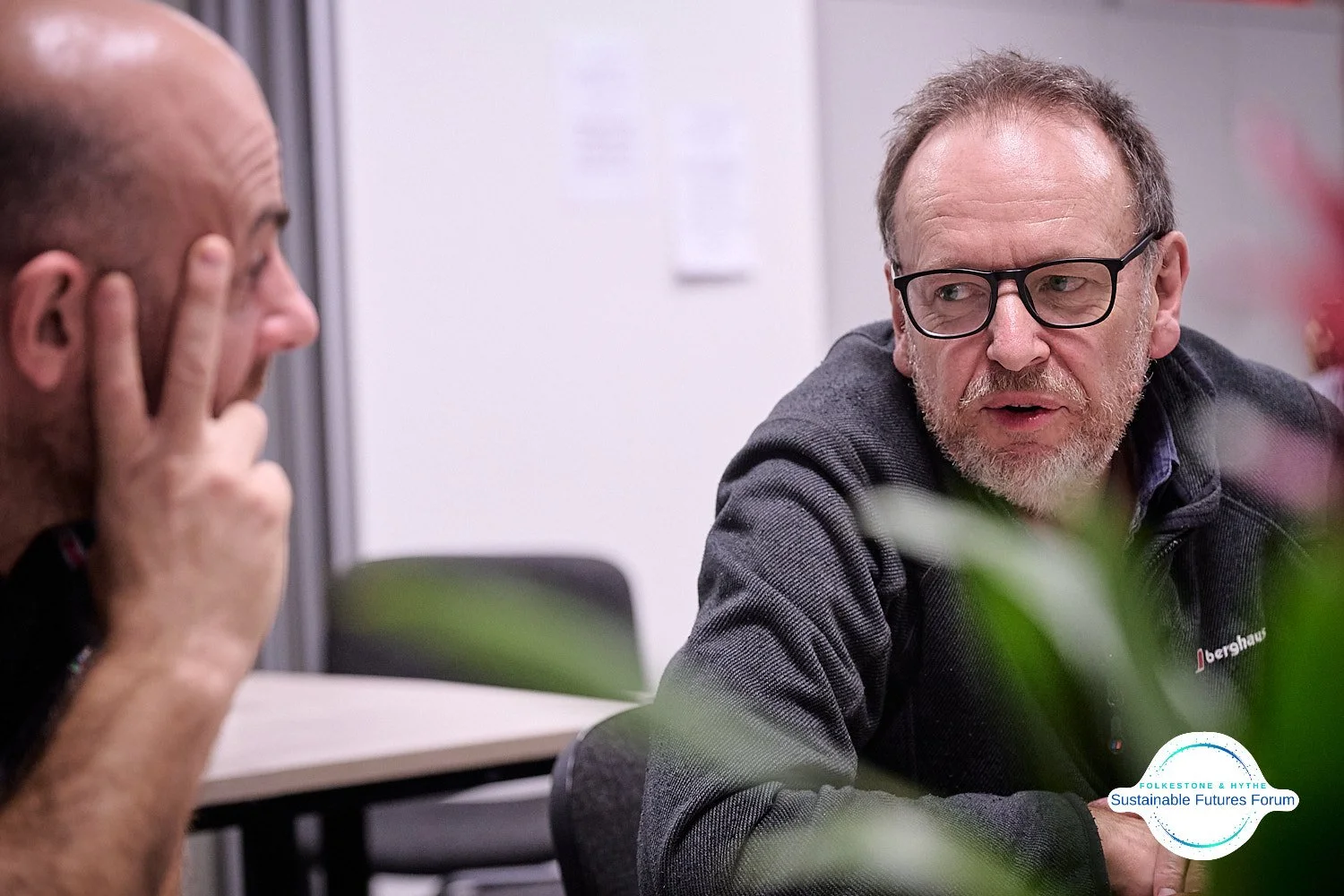 Two men having a conversation in an indoor setting, one with glasses and a gray beard, the other with a shaved head, with a pile of papers posted on the wall behind them. A logo for Folkestone & Hythe Sustainable Futures Forum is visible in the botto