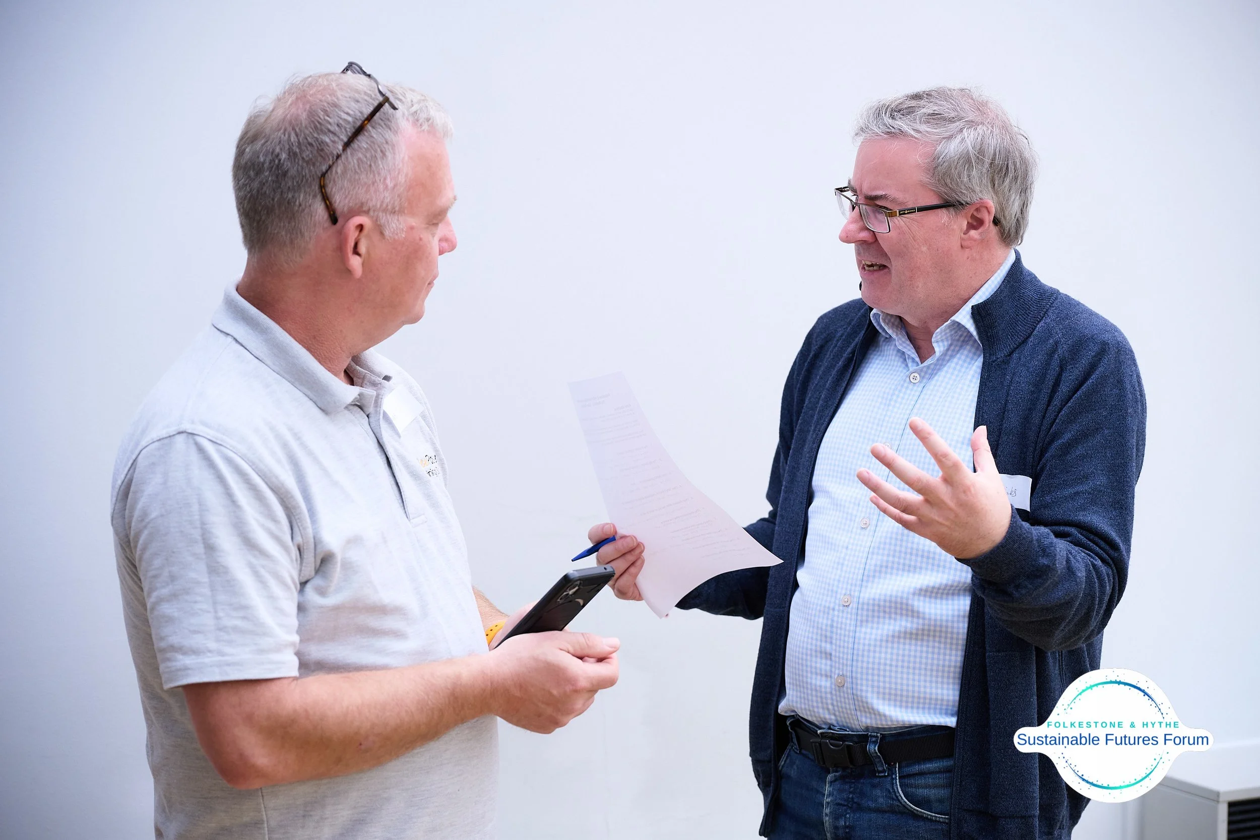 Two men engaged in conversation, one holding a smartphone and the other holding a paper, in a meeting or discussion setting, with a logo for Folkes Tone & Hythe Sustainable Futures Forum in the corner.
