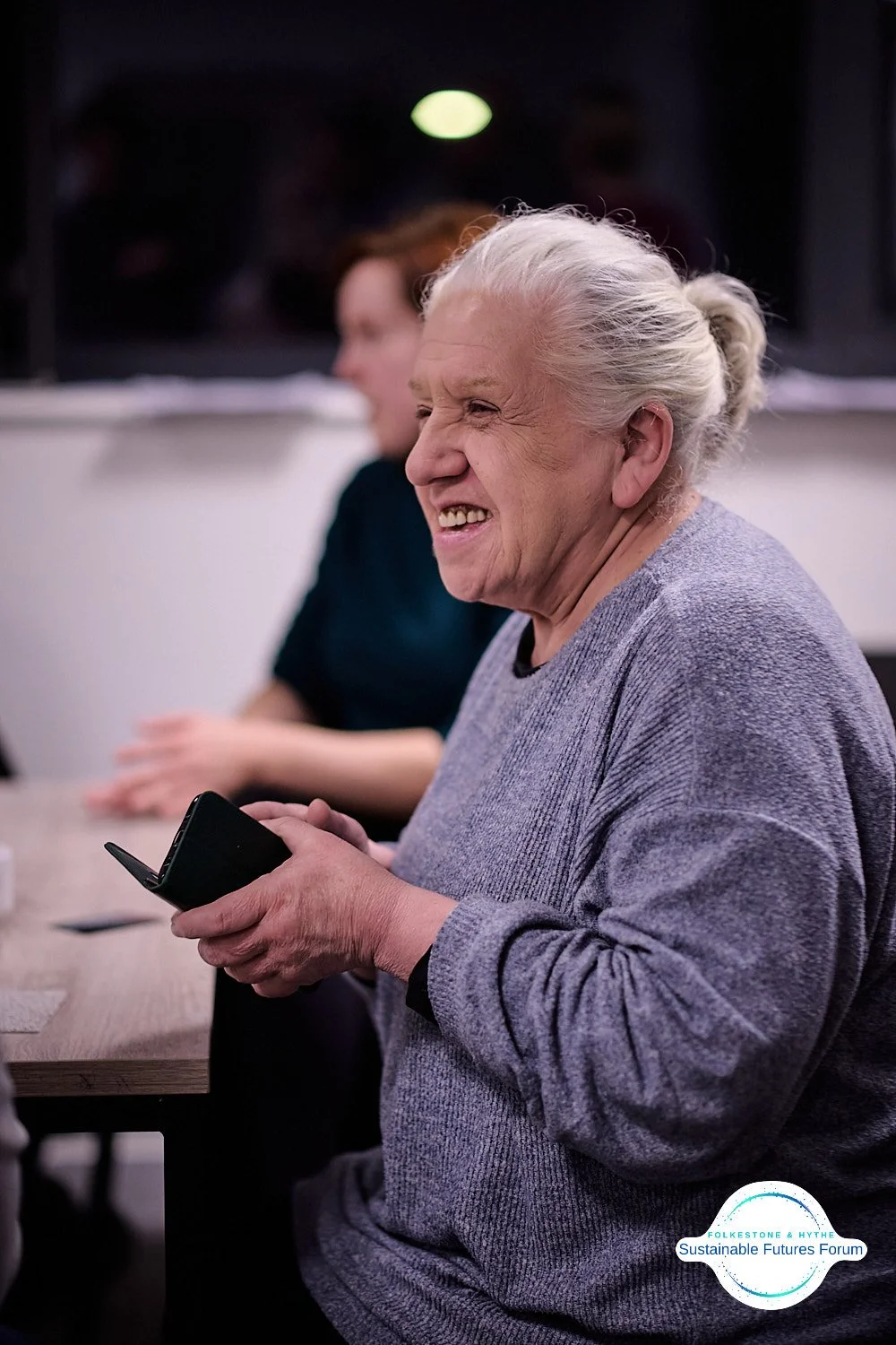 An elderly woman with gray hair tied back, laughing while holding a smartphone, sitting at a table with another woman blurred in the background.