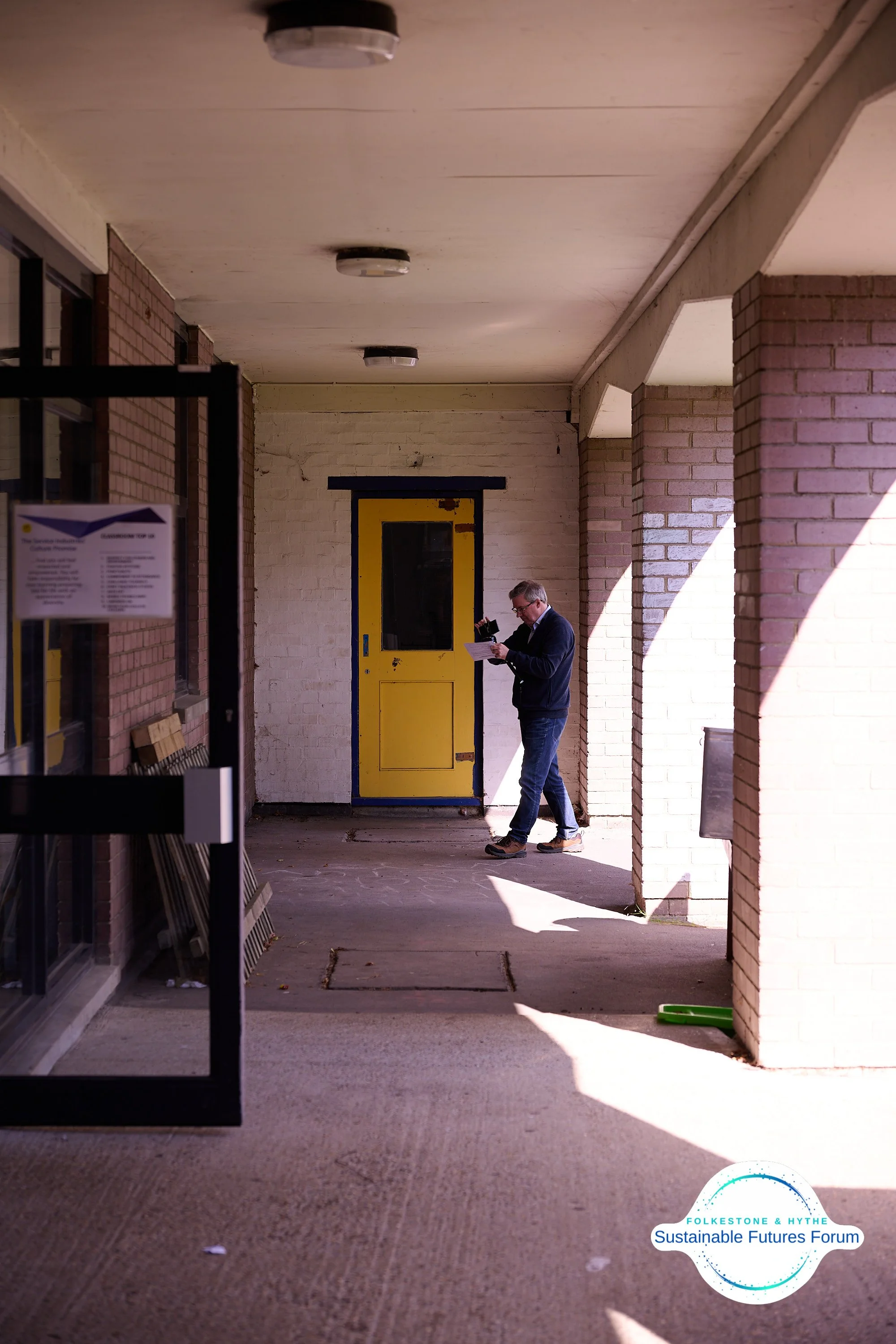 A man stands in a covered outdoor walkway of a brick building, reading a paper and speaking into a cellphone, with a yellow door behind him and sunlight casting shadows on the brick columns.
