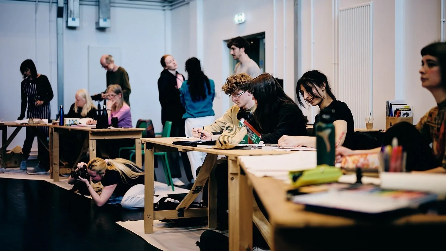 People sitting at desks in a classroom or meeting room, some working and others conversing, with a photographer kneeling on the floor taking pictures.