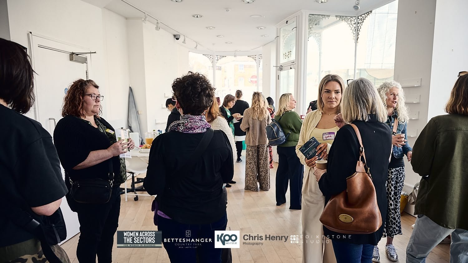 Women attending a networking event or meeting in a bright, modern room with large windows, holding cups and brochures, engaged in conversations.