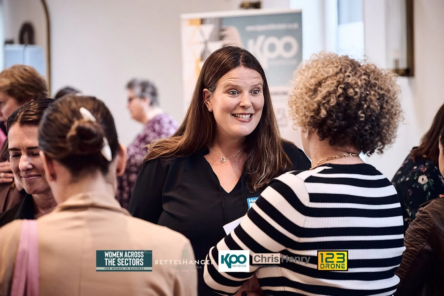 Women networking at a conference, engaging in conversations, with a woman smiling at a woman with curly hair in a striped top, in a room with others in the background.