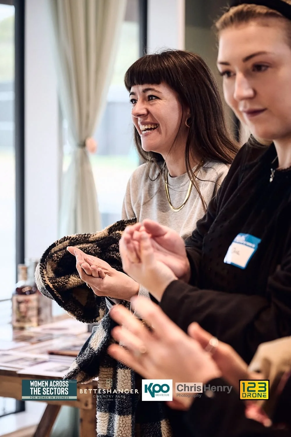 Two women talking at a women in business event, one smiling and wearing a beige sweatshirt, the other with a black jacket and a name badge.